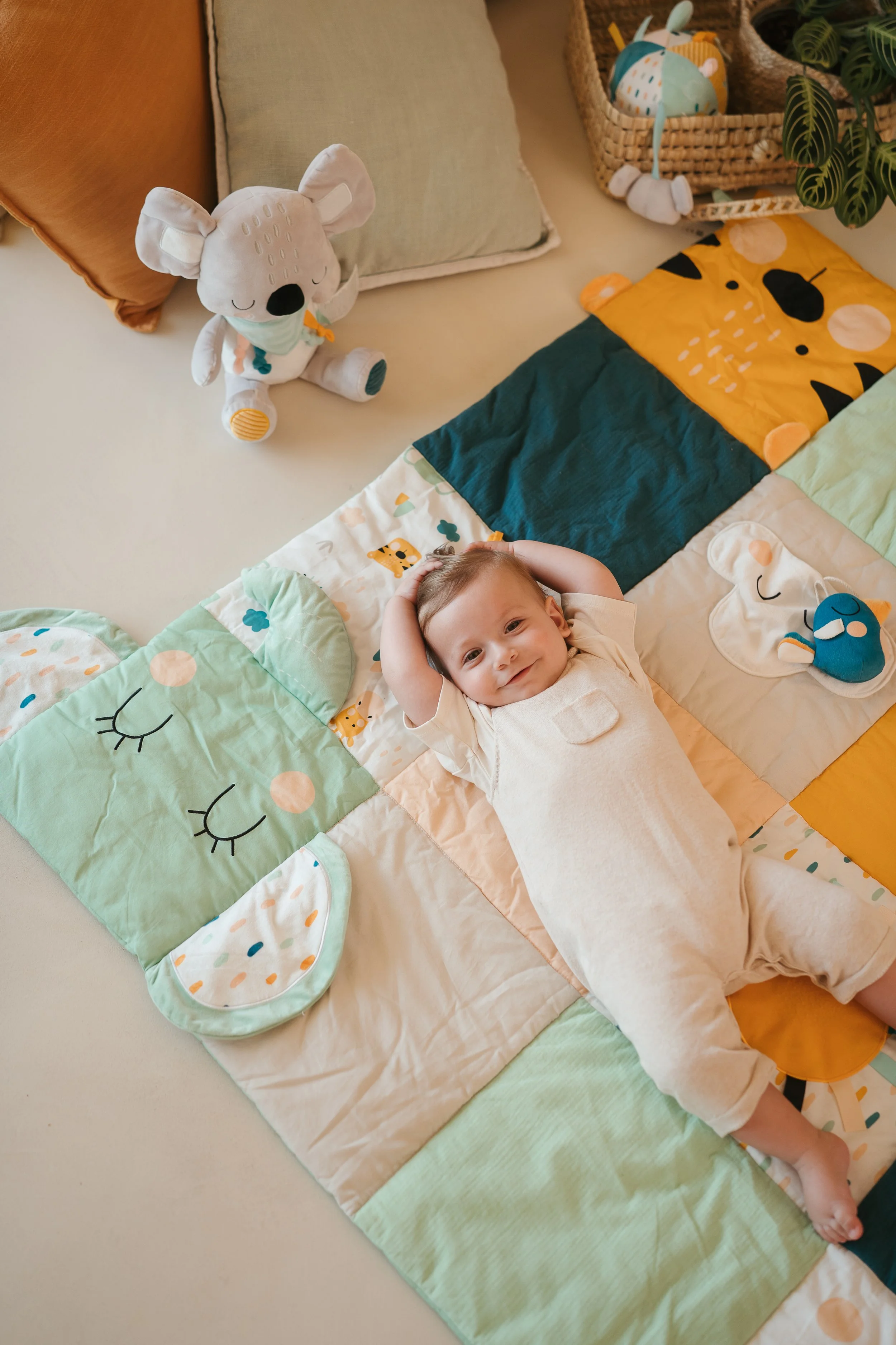 A young child lying on a colorful quilt with animal designs, smiling with hands behind head, surrounded by plush toys and cushions.