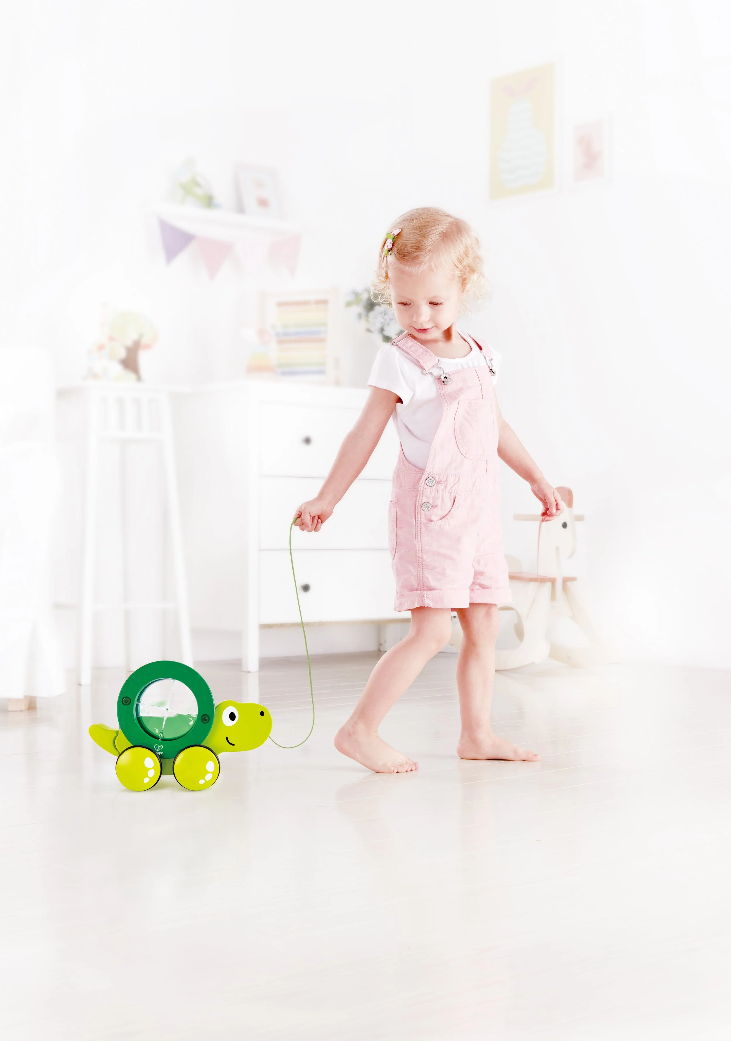 A young girl playing with a green and yellow turtle-shaped toy that has a clock on its shell, indoors in a bright, white room. Developed by Hape.