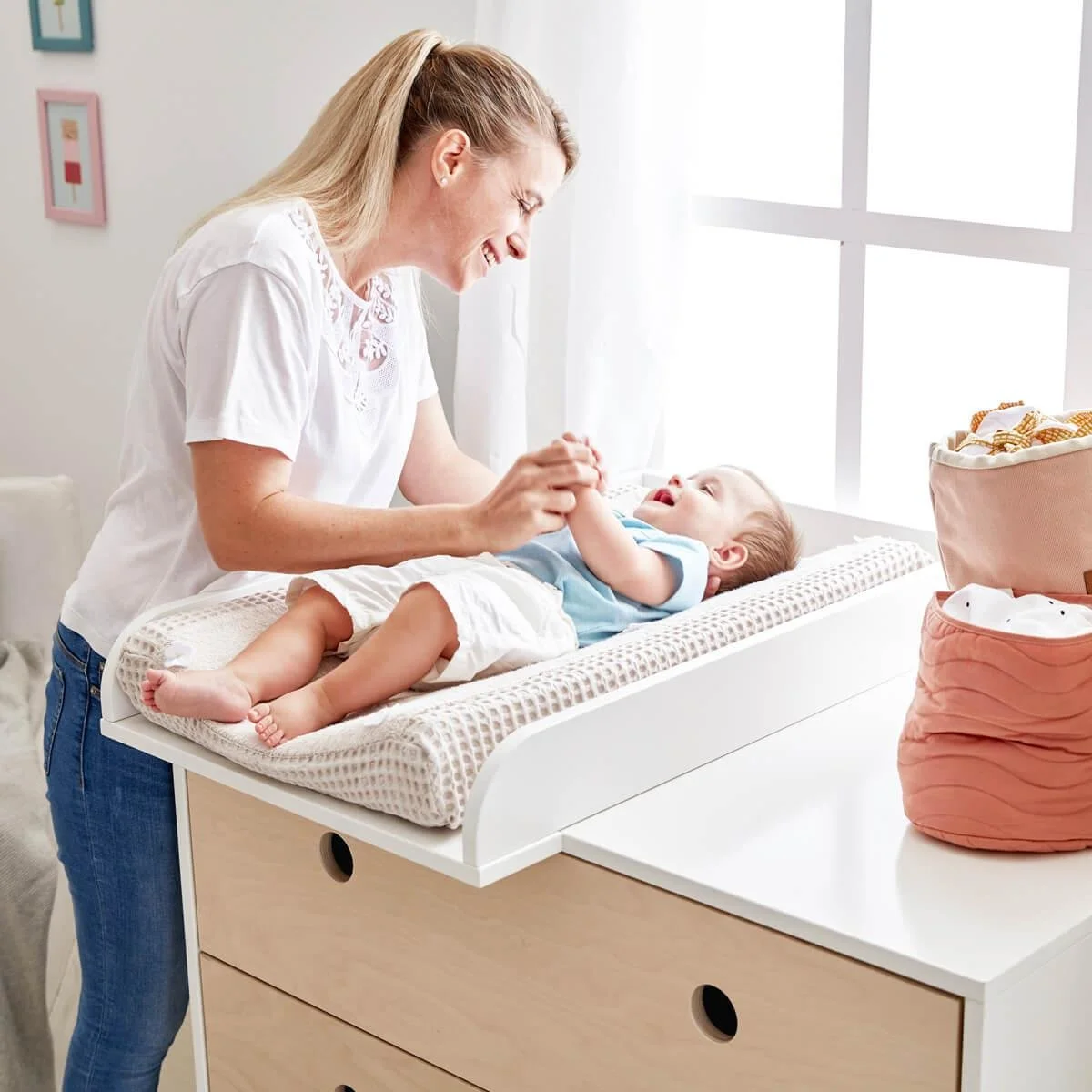 A woman with blonde hair smiling and holding a baby's hands while the baby is lying on a Colorflex changing table.
