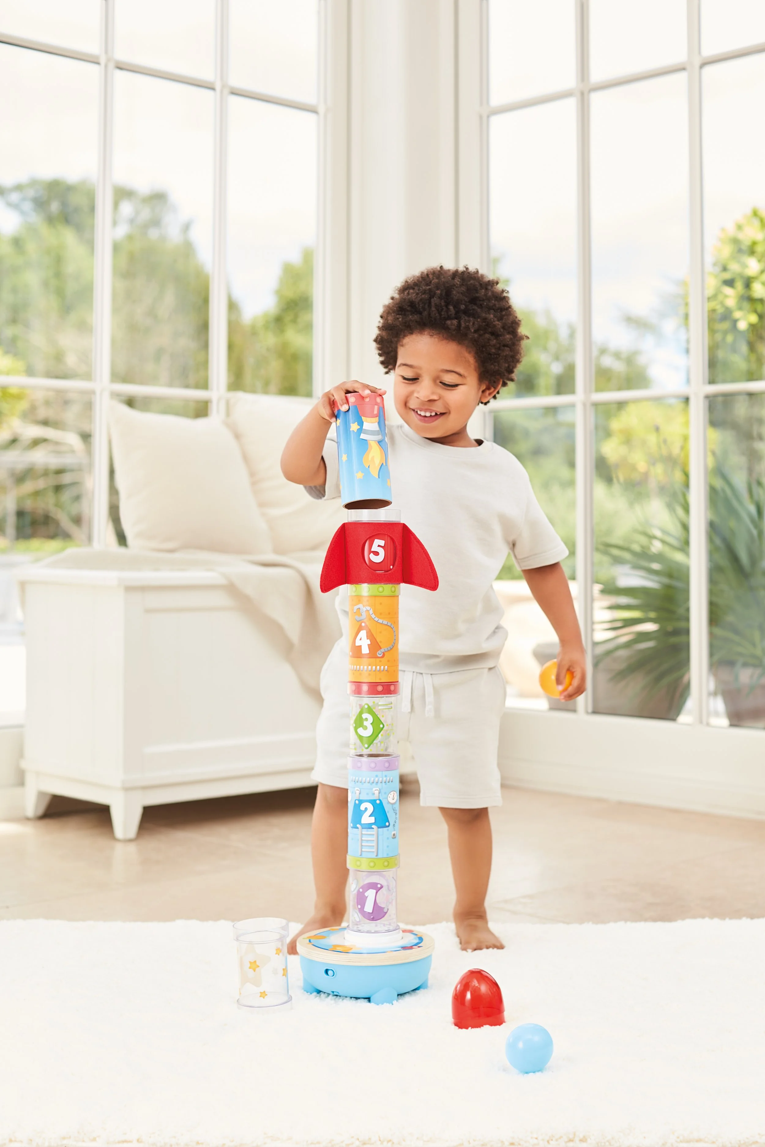 A young boy playing with a stacking rocket toy, stacking parts from 1 to 5, in a sunlit room with large windows and white furniture.