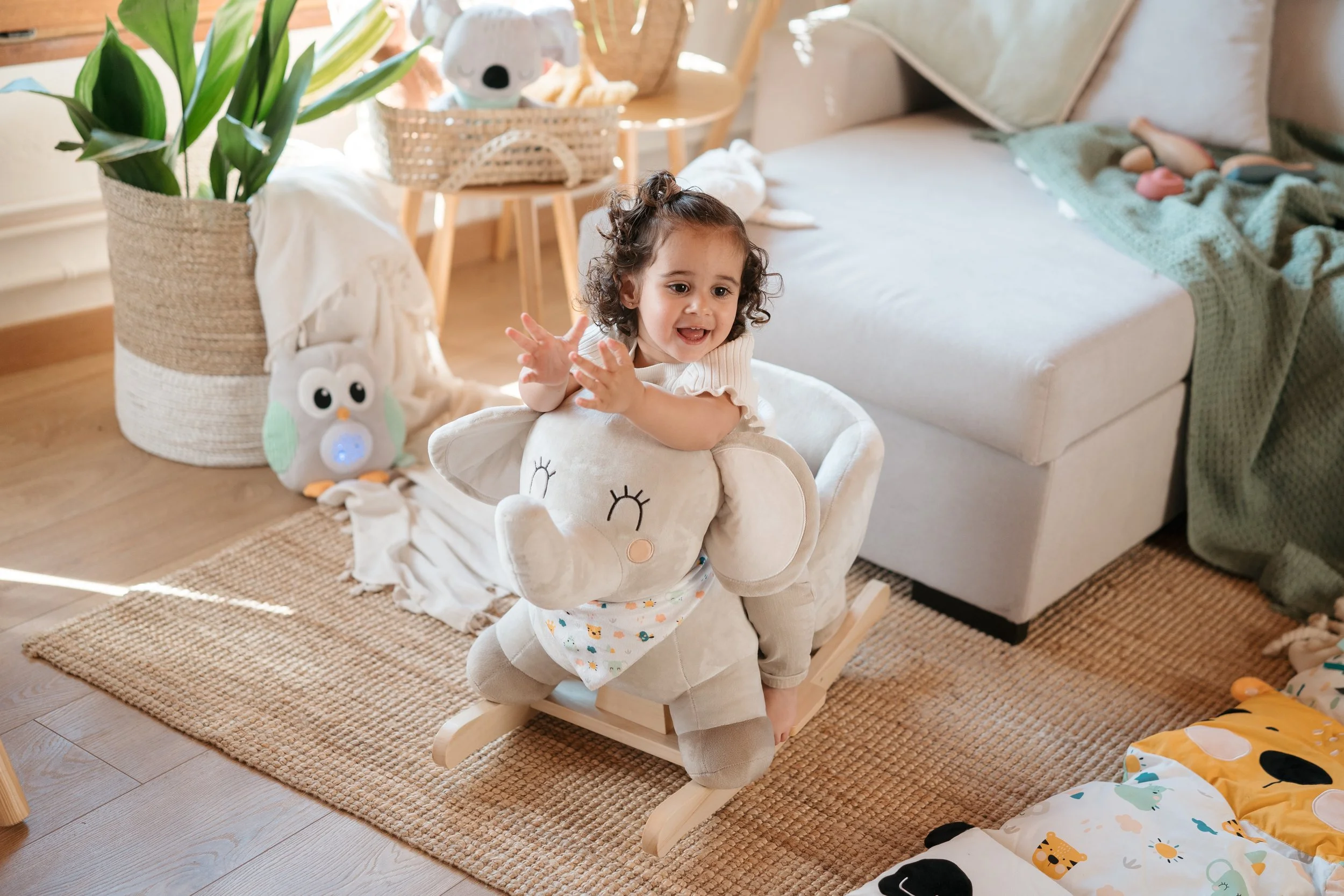 Young girl sitting on a plush rocking elephant toy in a cozy living room, surrounded by toys and accessories from the Eurekakids Cucu collection.