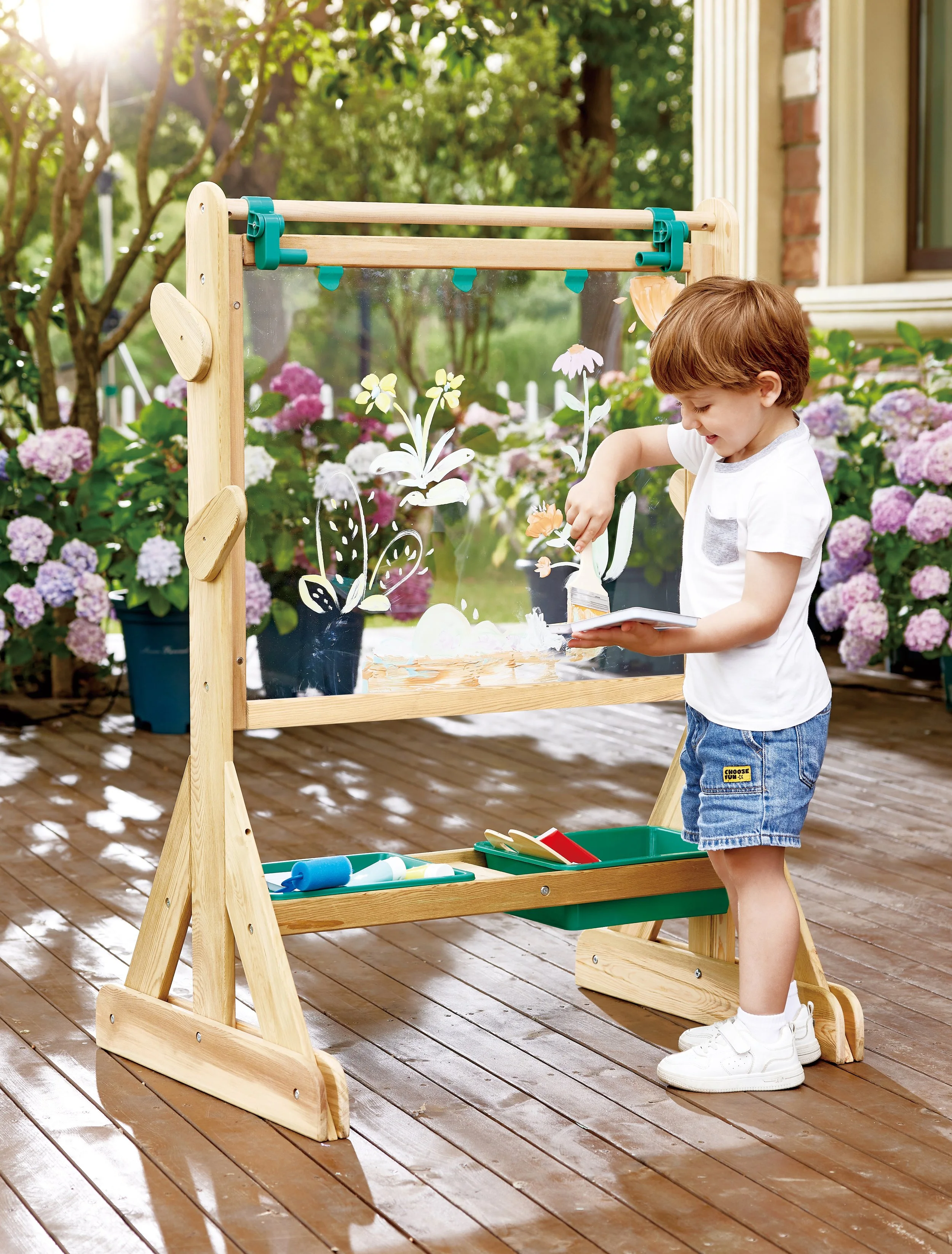 A young boy painting and drawing on the Hape outdoor easel on a wooden patio, with pink and purple hydrangea flowers and green trees in the background.