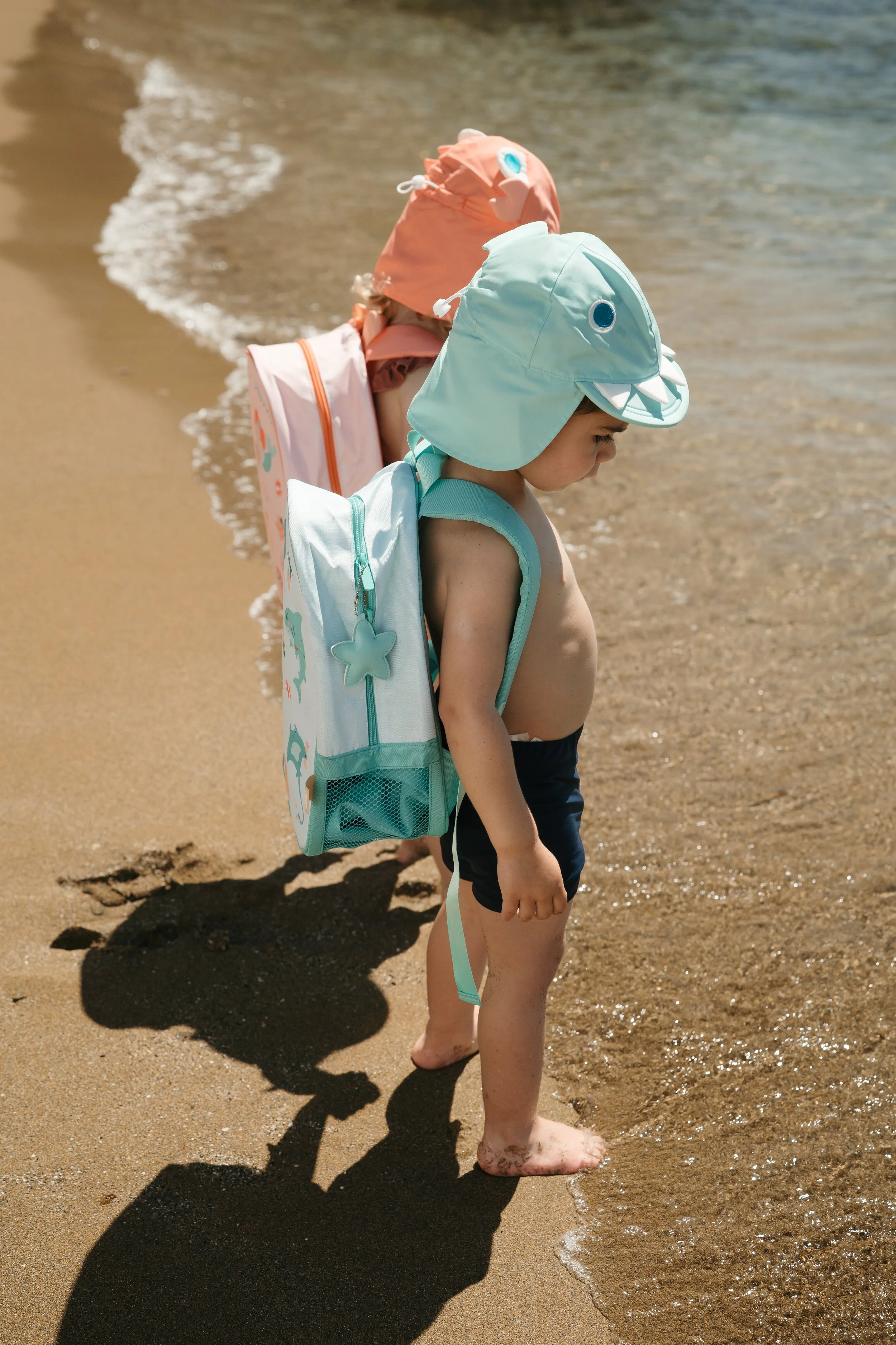 Young child at the beach wearing a shark-themed hat from the Eurekakids Beach Collection and carrying a matching backpack, standing barefoot on the sand near the shoreline with waves.