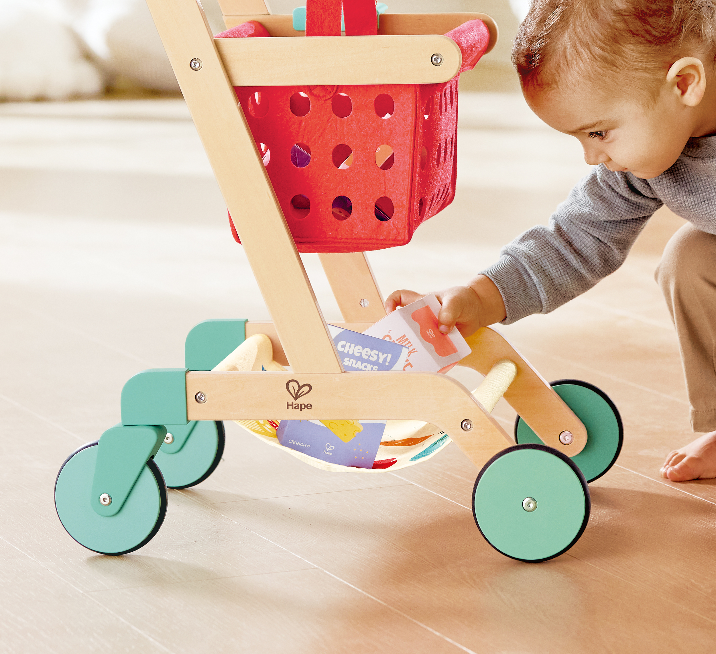 Young child placing snack item into toy shopping cart with red basket and wooden frame on light-colored floor in a playroom.