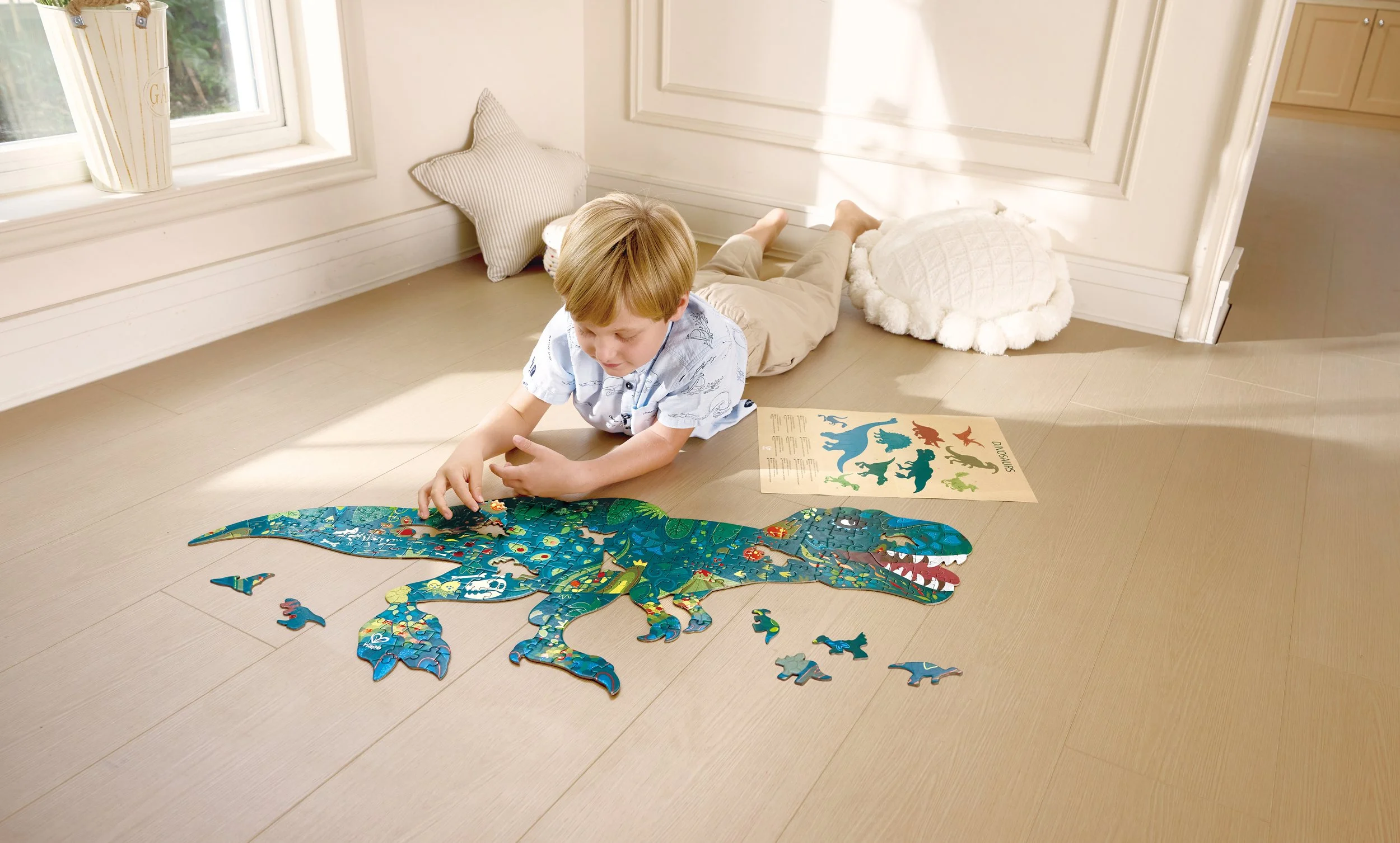 A young boy is assembling a Hape puzzle of a blue dinosaur on a light-colored wooden floor near a window, with some puzzle pieces and a dinosaur poster nearby.