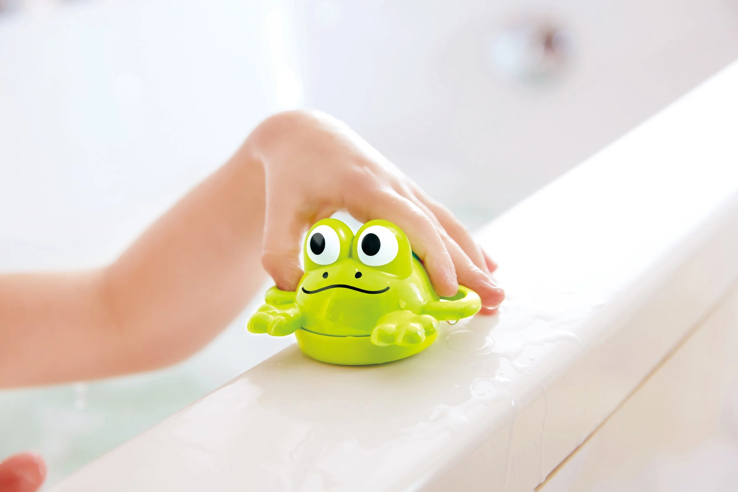 Child's hand holding a green frog-shaped bath toy that is part of "double Fun Fishing Set" by Hape, in a bathtub.