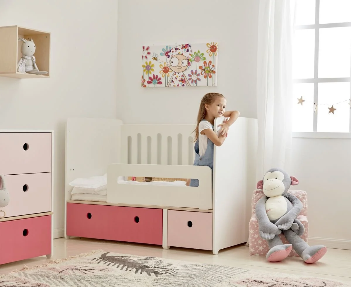 A young girl standing on a Colorflex bed in a nursery, smiling and gazing out the window. The room has pastel-colored furniture, a stuffed monkey toy sitting on a pink box, and wall decorations including a colorful artwork and star-shaped garland.