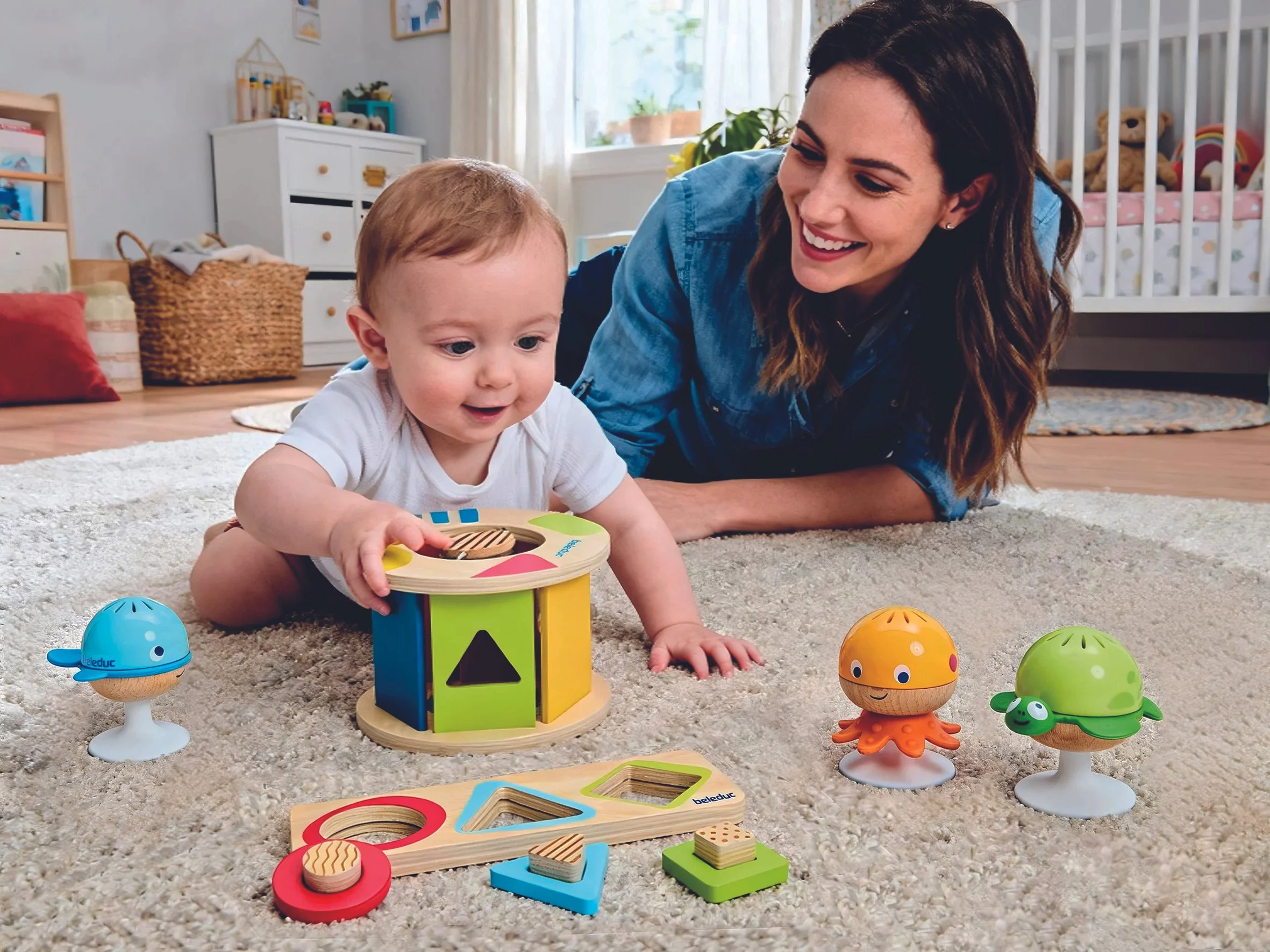 A baby and a woman playing with colorful wooden toys on a beige carpet in a bright, cozy nursery. The baby is holding a circular toy, and there are various animal-shaped and geometric toys around them.