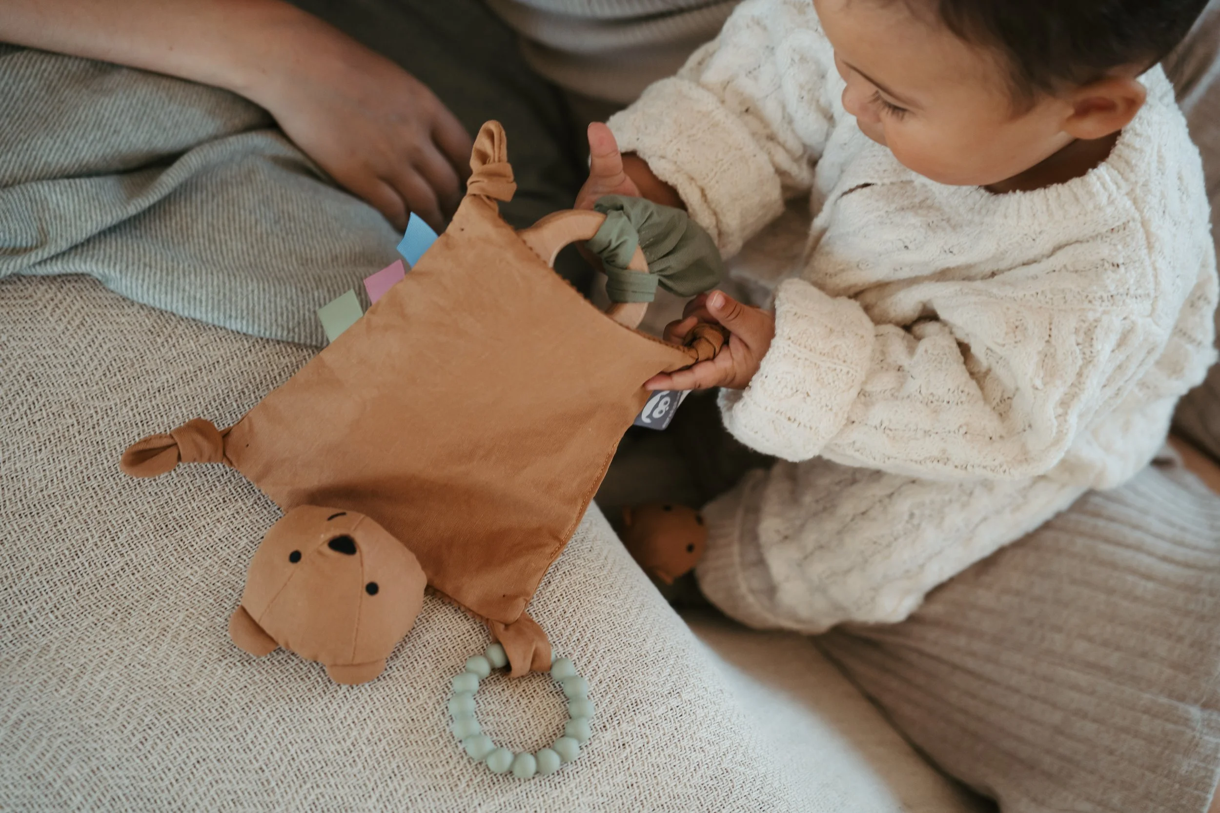 A young child in a cream-colored sweater playing with a soft brown plush toy that has a stuffed bear face and a beaded teething ring attached, sitting on a beige couch.