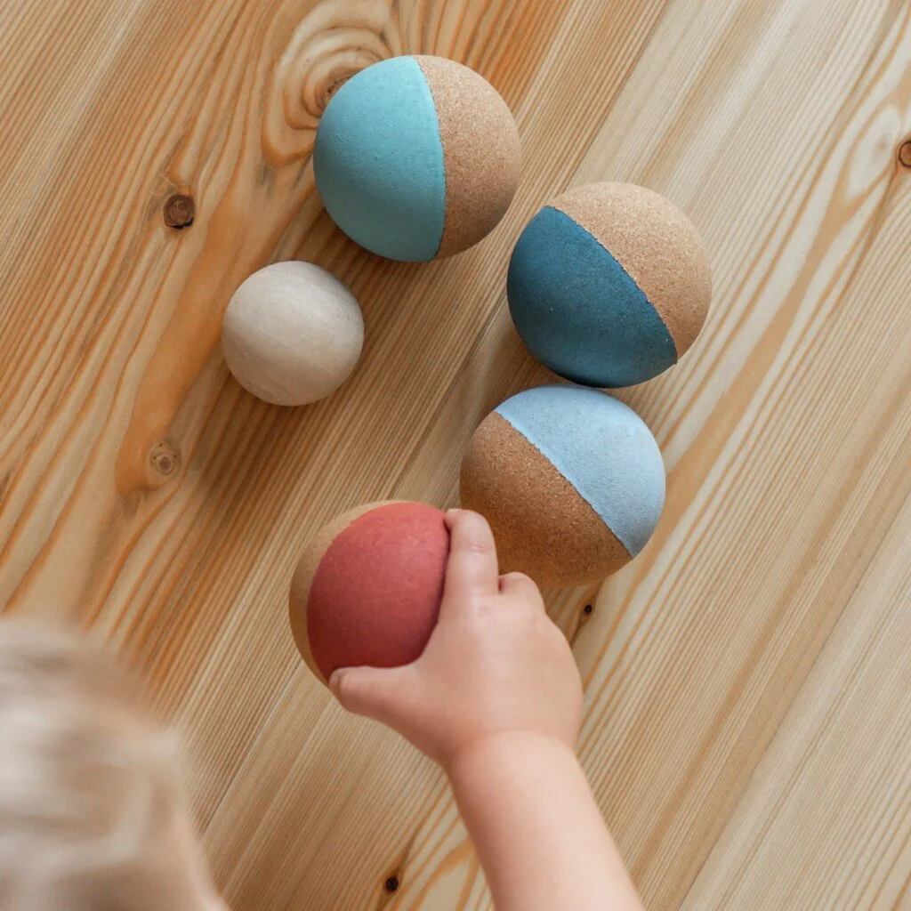 Child's hand reaching to pick up a red and pink half-sphere, with several geometric shaped balls, some remain uncolored, some have blue and beige painted halves, placed on a wooden surface. By Korko.