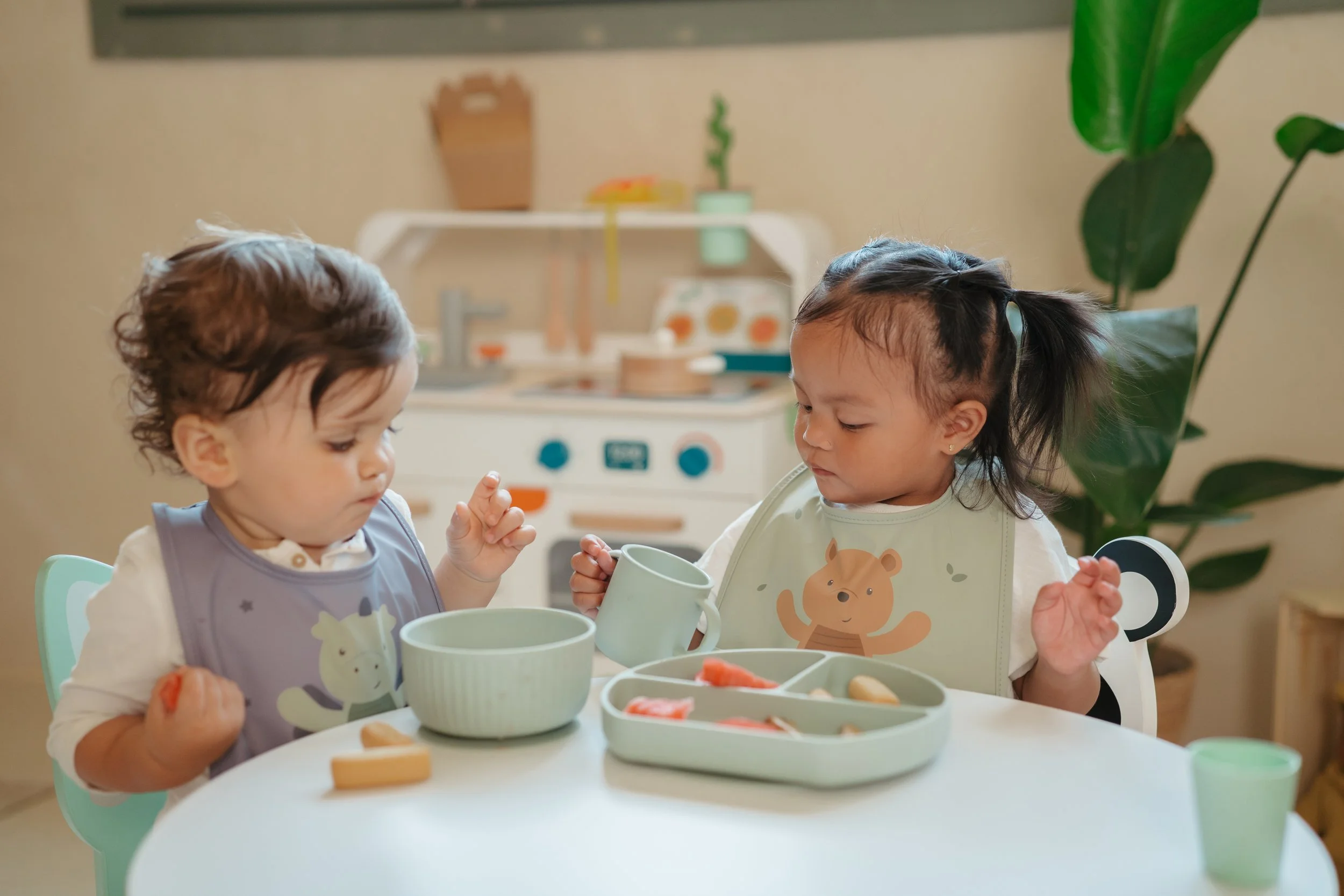 Two young children sitting at a white table with pastel-colored dishes and cups, playing with toy food in a cozy, well-lit playroom with a toy kitchen in the background and a large green plant.
