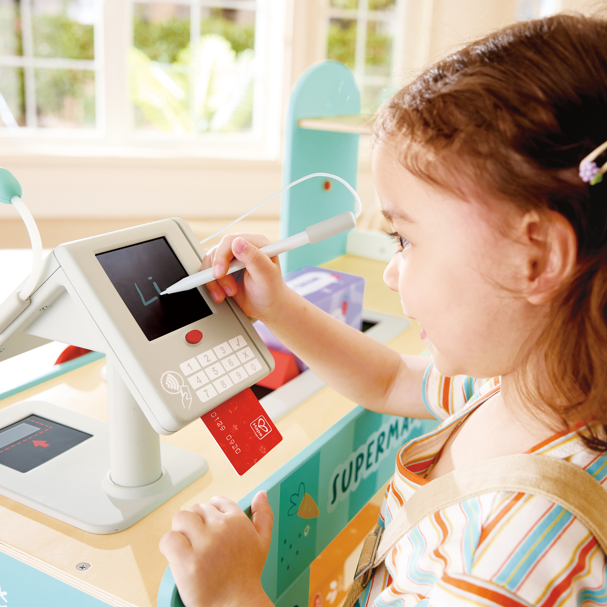Young girl using a stylus to write 'Li' on a small electronic device at a kids' play area with colorful decorations.