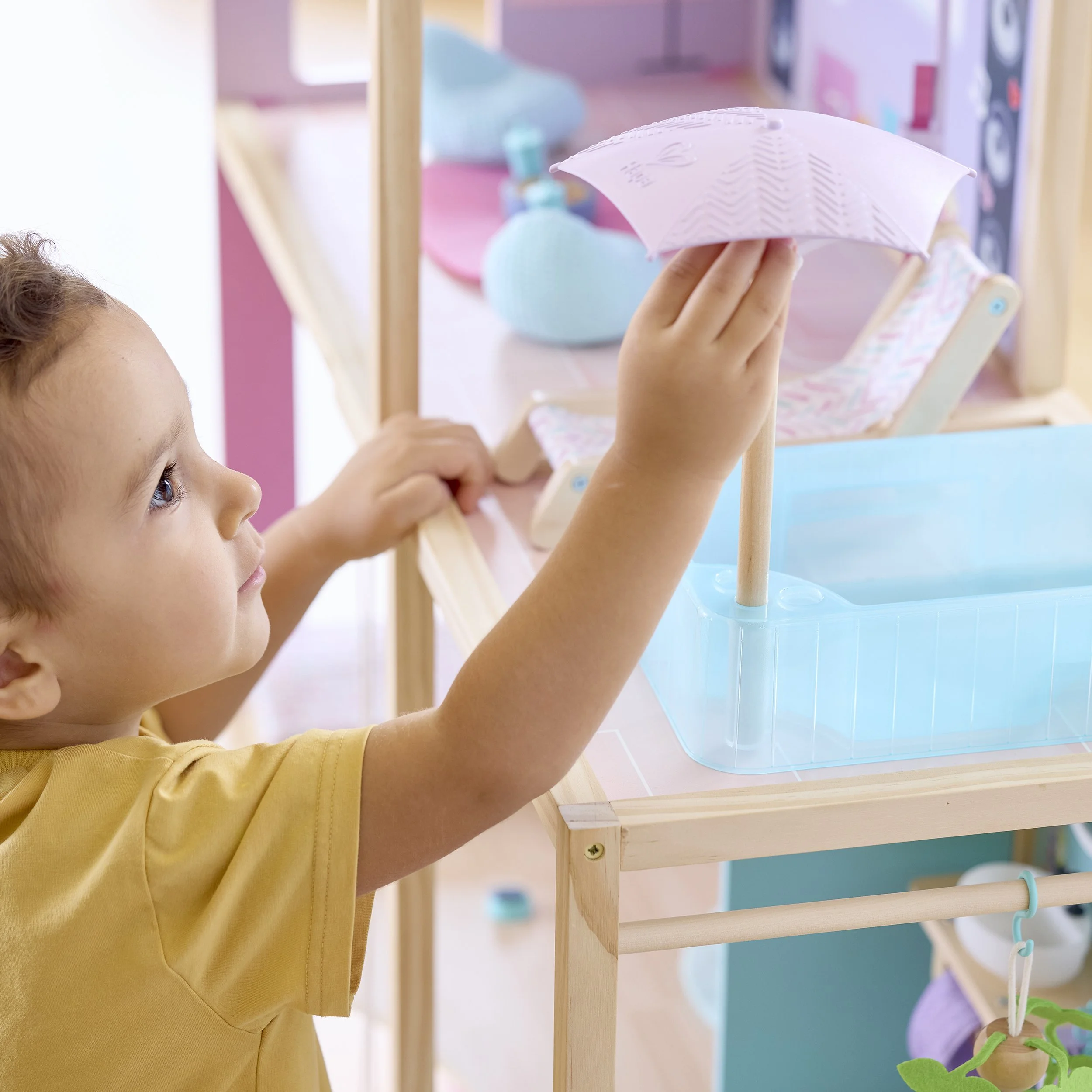 A young child with a yellow shirt playing with a dollhouse, holding a white umbrella and looking at the dollhouse.