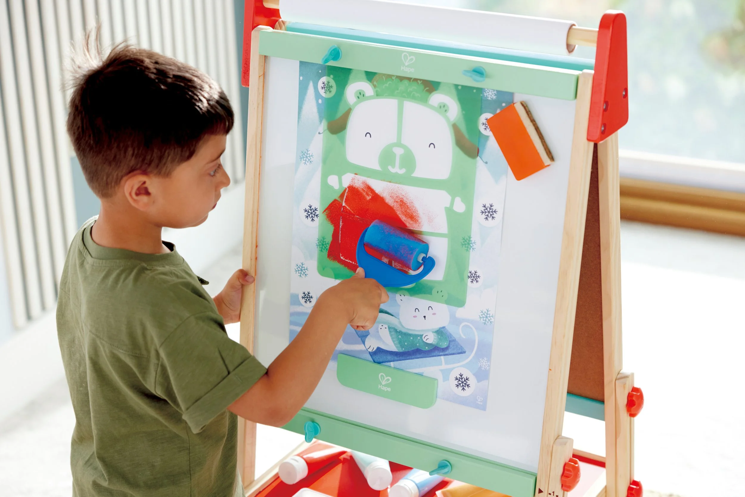A young boy with short brown hair is standing in front of the Hape colorful drawing board, using a blue roller to apply red paint on paper attached to the board with hooks. The drawing board features a cute cartoon bear with winter-themed decorations