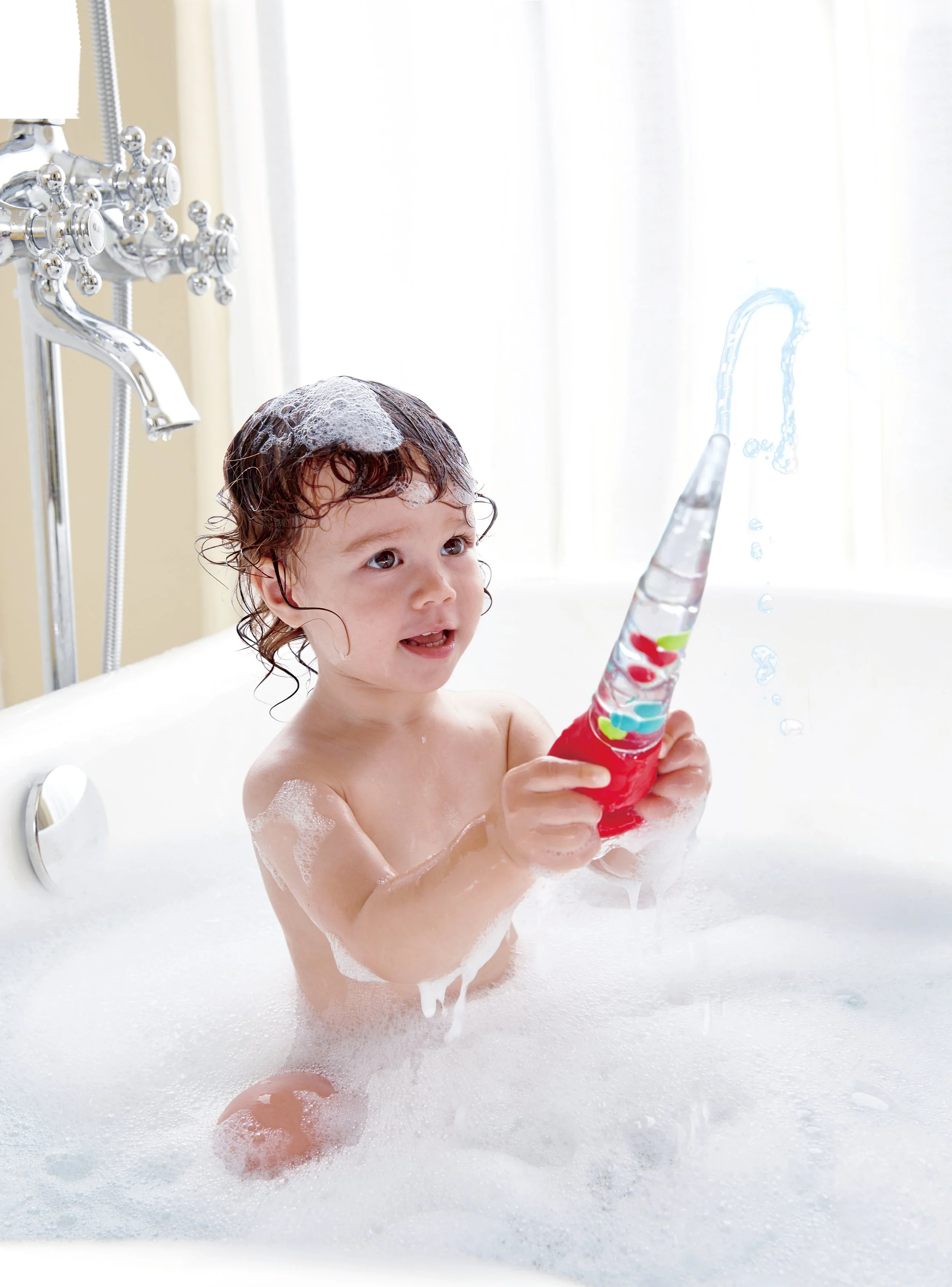 A young child playing with "Squeeze and Squirt Pipette" by Hape in a bubble-filled bathtub during bath time.