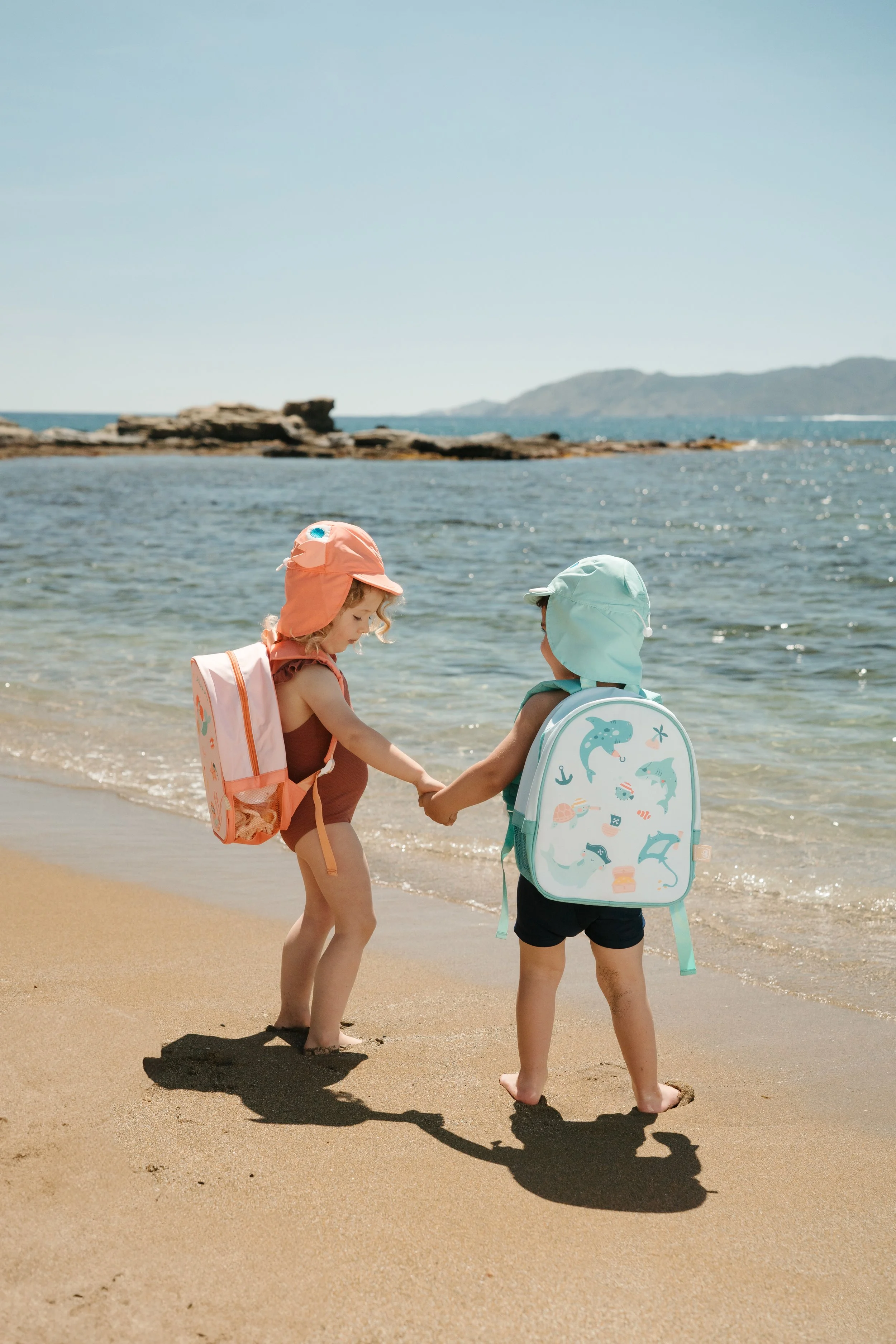 Two young children wearing backpacks from the Eurekakids Beach Collection, holding hands on a sandy beach near the water, with rocky formations and distant hills in the background.