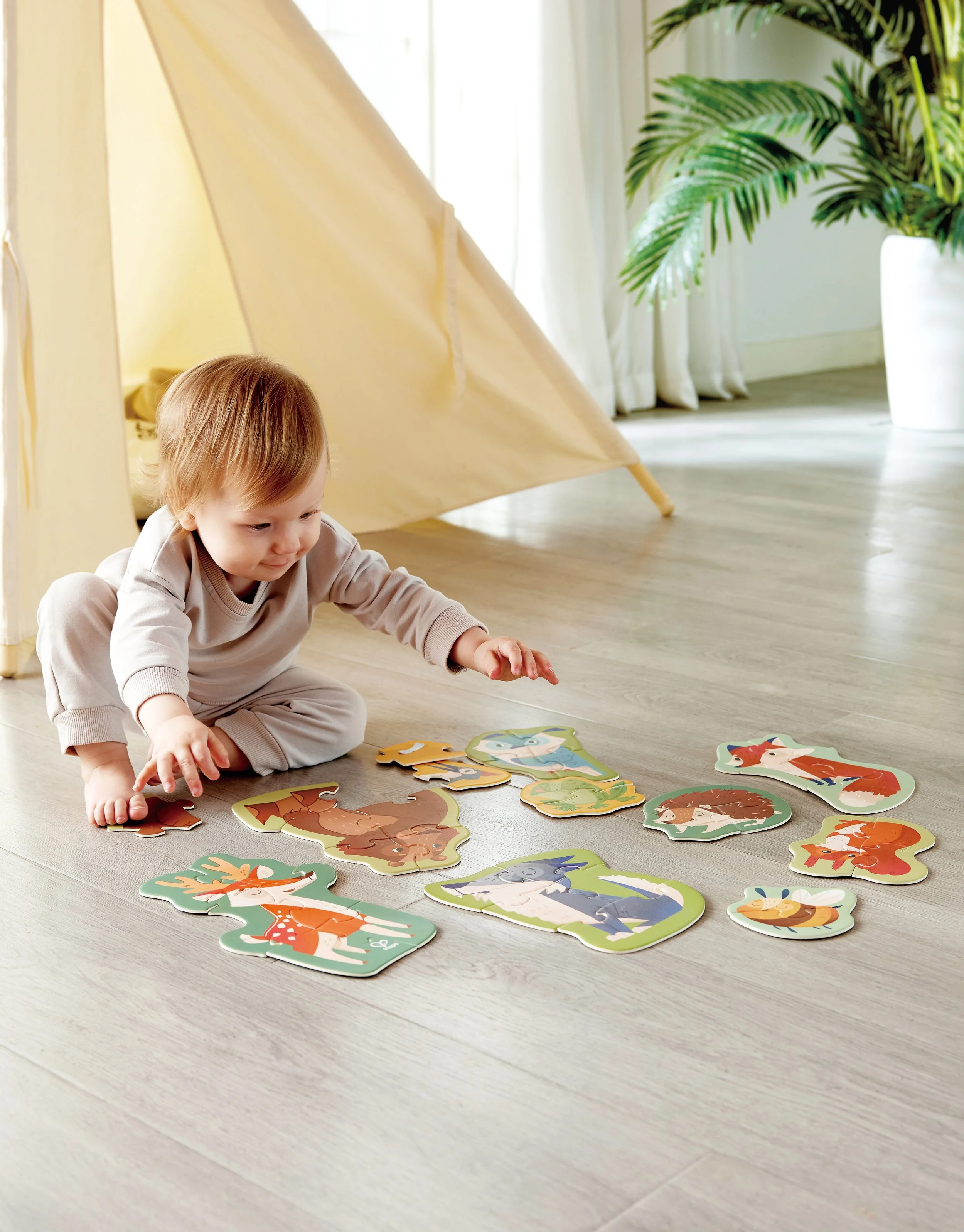 A young child playing with animal-shaped Hape puzzle pieces on a hardwood floor in a room with a tent and large potted plant.
