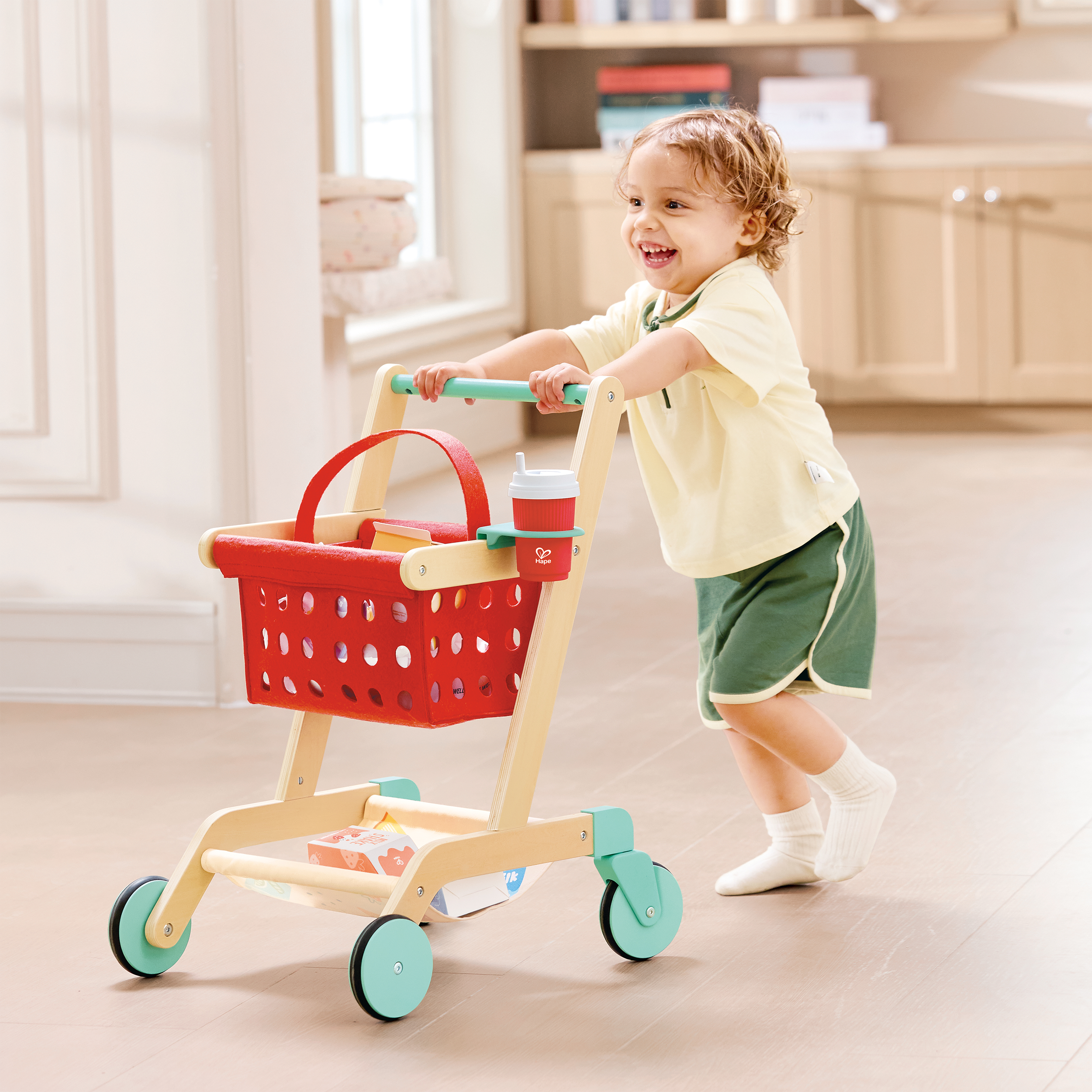 A young child with curly hair, wearing a light yellow shirt and green shorts, is pushing a toy shopping cart inside a bright kitchen.