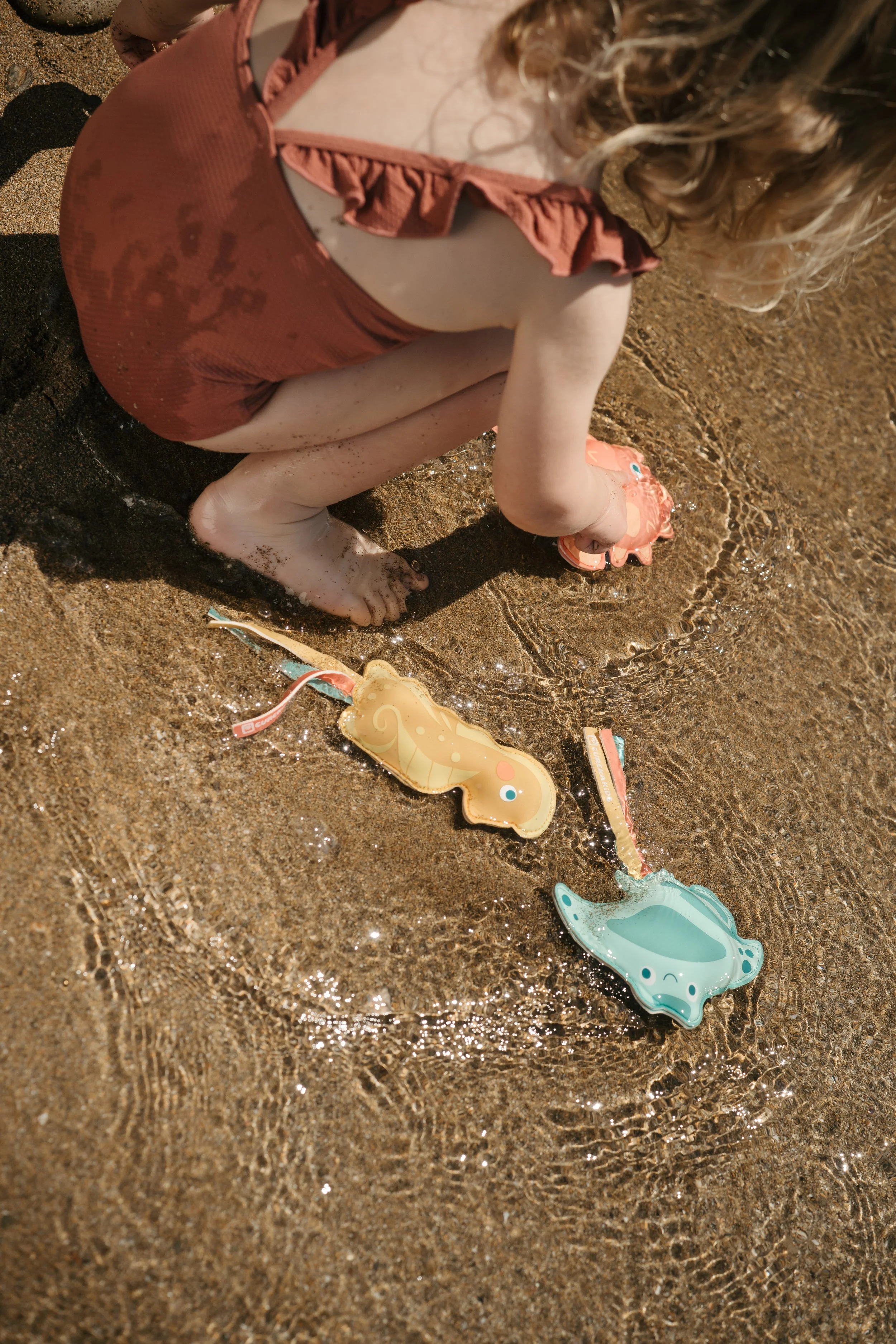 A young girl in a tan swimsuit playing in shallow water at the beach, with bath toys from the Eurekakids Beach Collection.