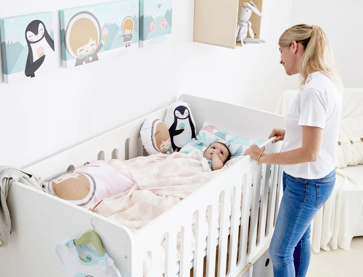 A woman leaning over a white Colorflex crib, looking at a baby lying in bed. The crib has pillows with penguin and girl faces, and there are colorful children's paintings on the wall.
