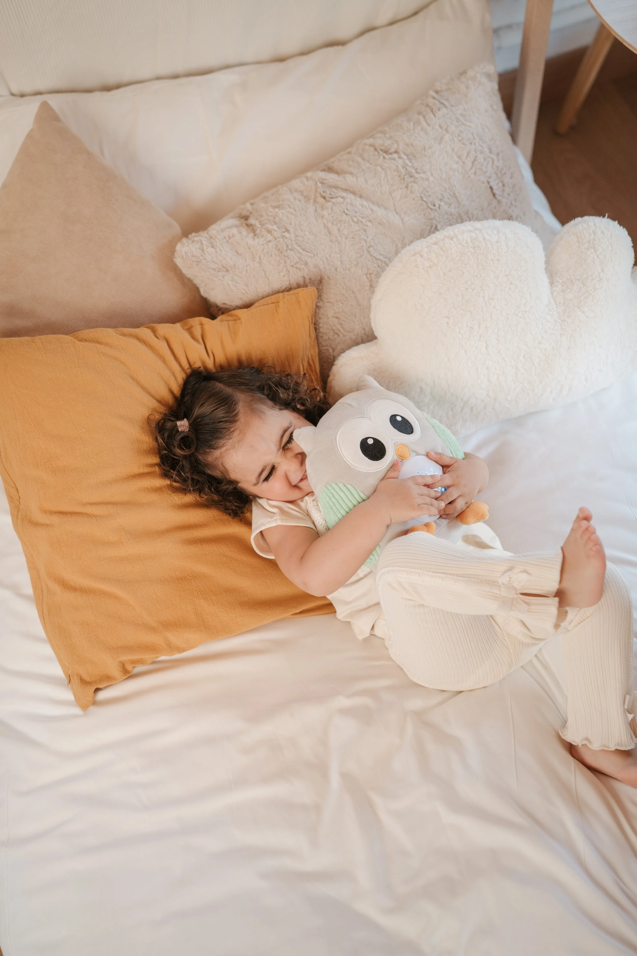 Young girl lying on a bed, hugging a Archi plush from Eurekakids Cucu, surrounded by fluffy pillows.