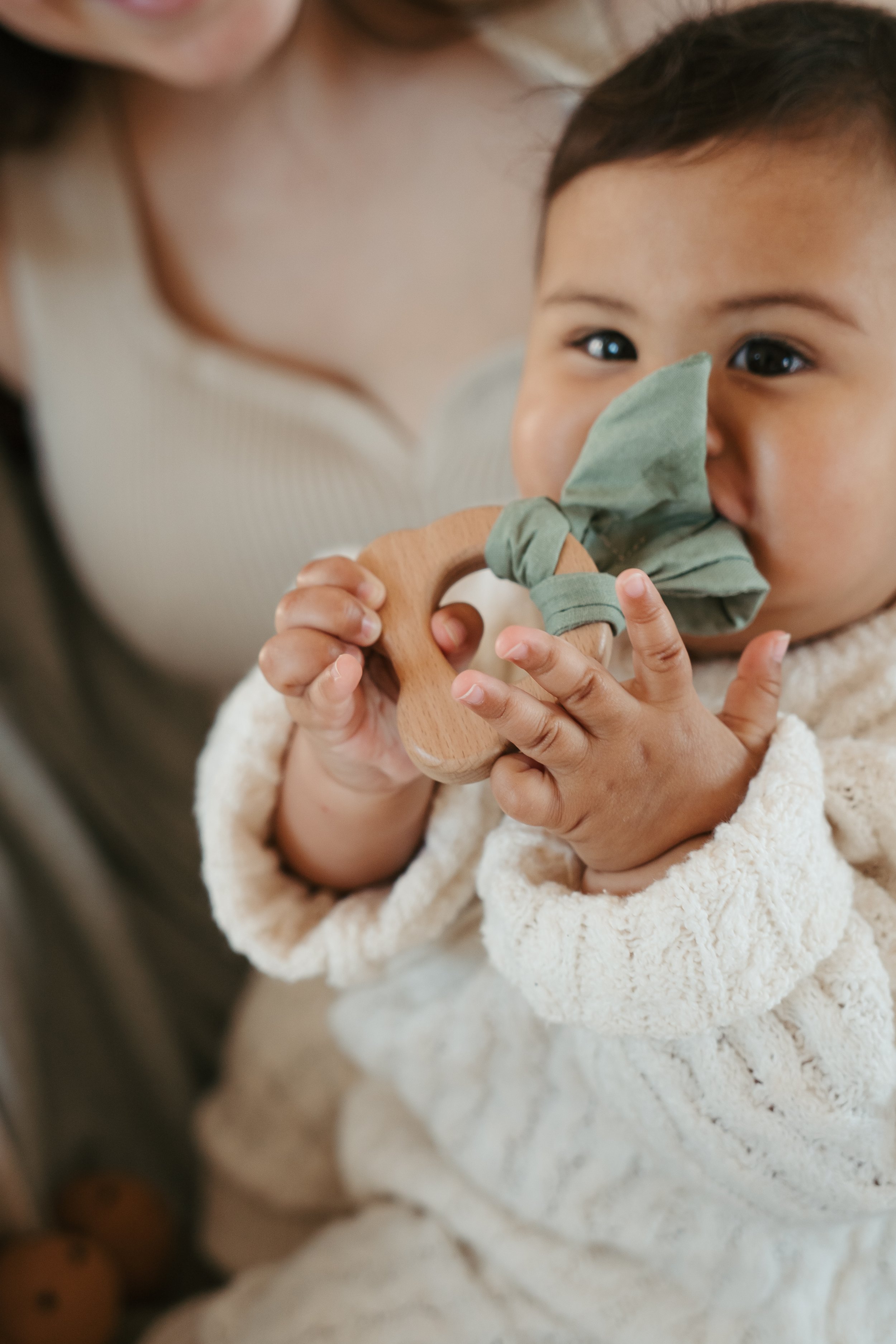 A young Asian child holding a wooden teething toy with a cloth tied around it, touching the cloth with their nose, while sitting on a person's lap. The child is wearing a cream-colored cozy sweater.