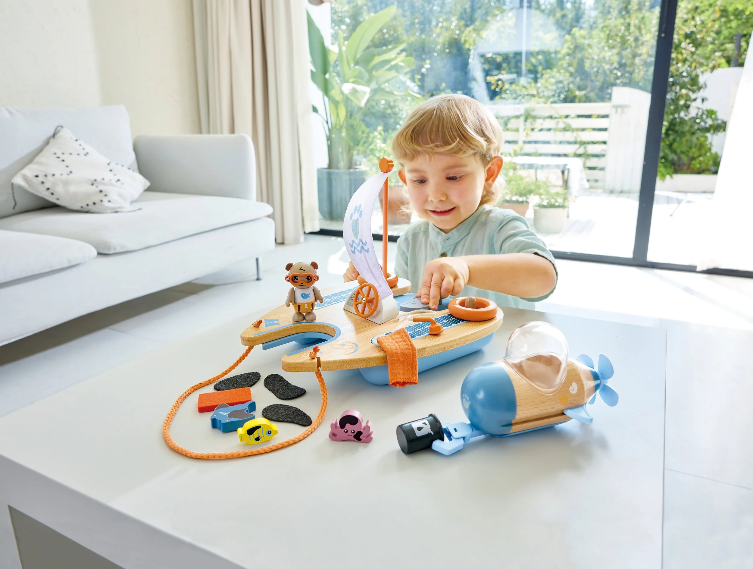 A young boy playing the Green Planet Explorers Ben’s Eco Catamaran playset by Hape, a wooden toy boat with a sail on a white table in a bright living room.