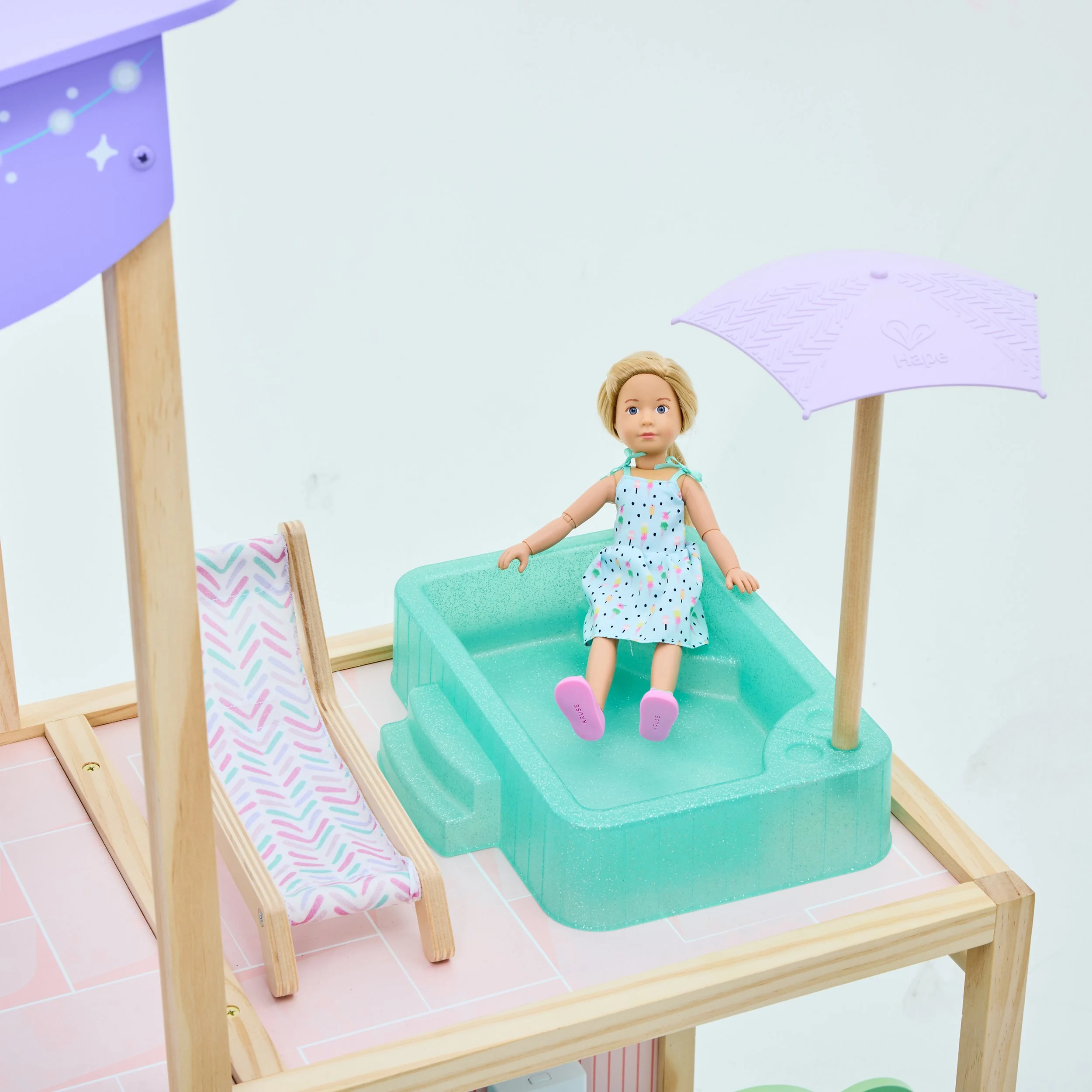 A doll sitting in a small, teal plastic pool with a purple umbrella and a pink and white patterned lounge chair nearby on a wooden platform.