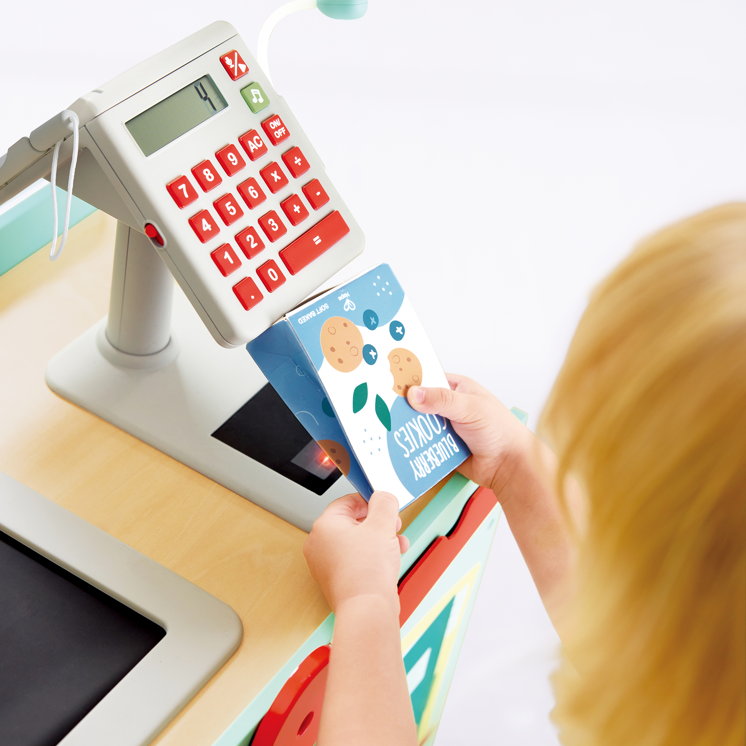 A child using a toy checkout register with a calculator, scanning a blue box labeled 'Blueberry Cookies' at a play store or pretend shop setup.