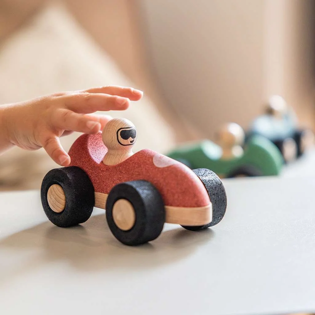 Child's hand reaching to touch a wooden toy race car with a smiling face design. By Korko.