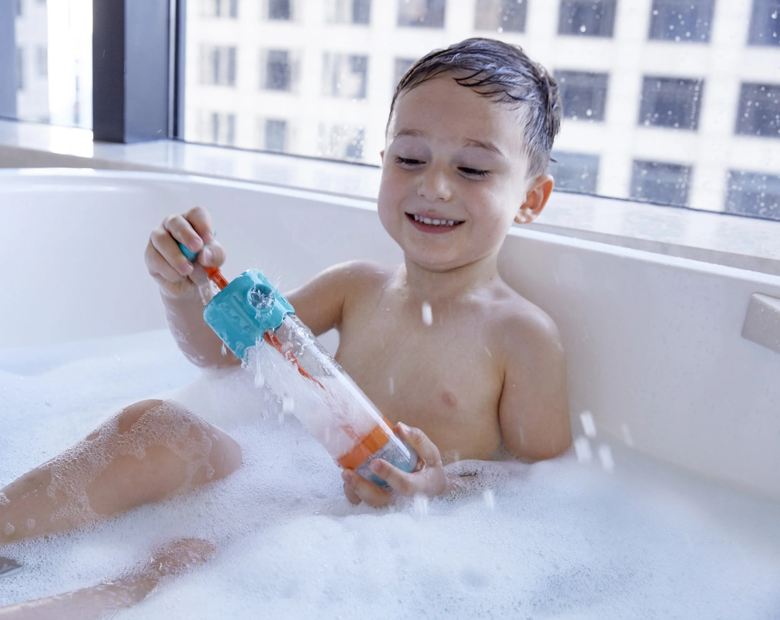 A young boy playing with "Multi-spout sprayer" by Hape in a bubble-filled bathtub, smiling with wet hair, in an urban high-rise bathroom with city buildings visible through the window.