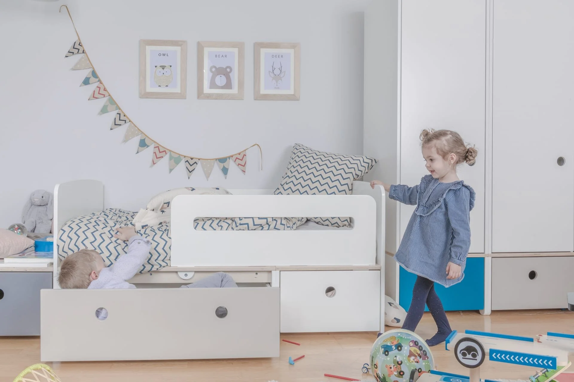 A young girl in a blue dress is standing near a white Colorflex bed with a little boy lying on it in a bedroom. Decorations include framed animal prints, a colorful bunting, and toys scattered on the floor.