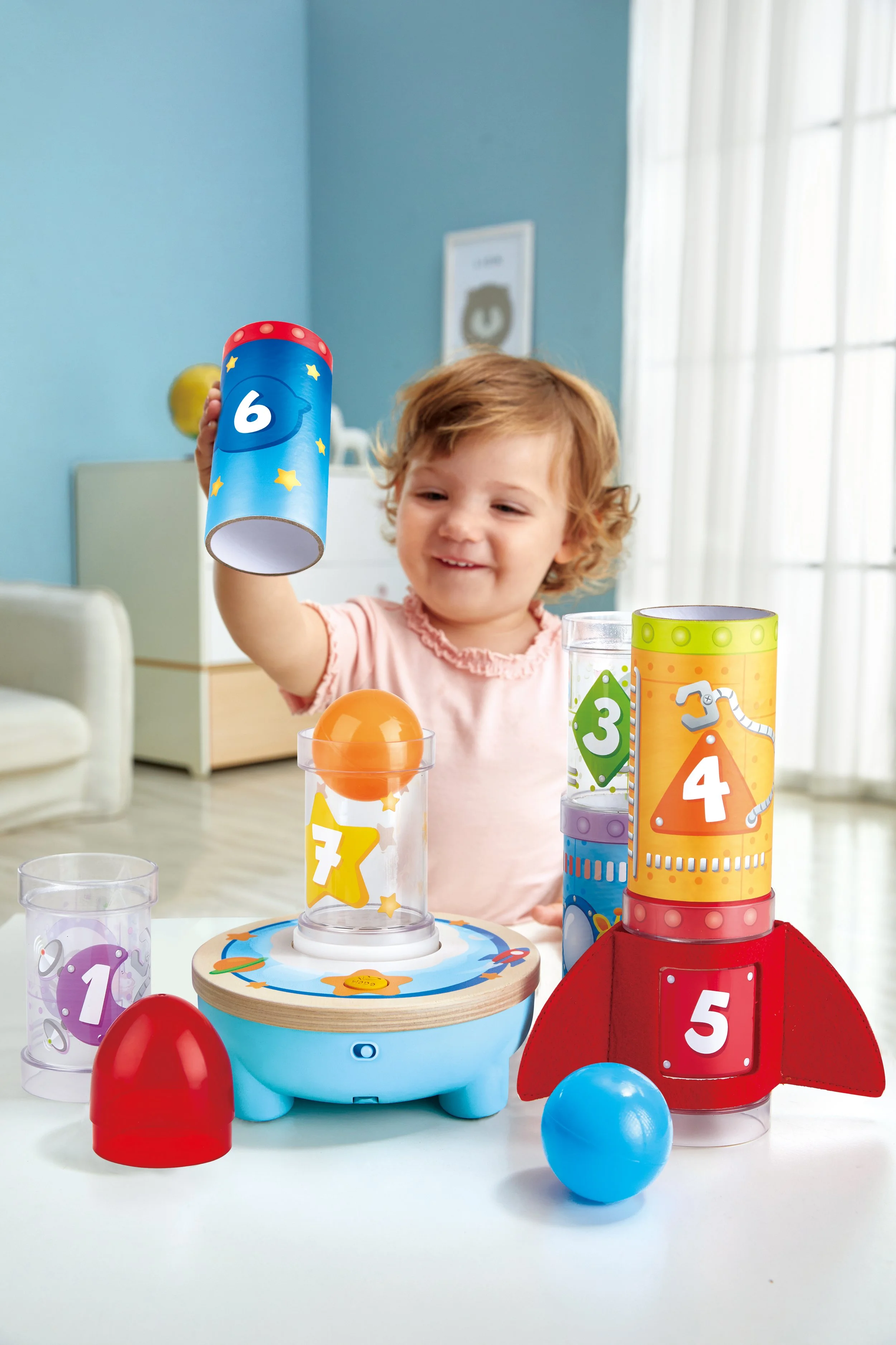 A young child playing with colorful stacking and sorting toys, including a rocket-shaped toy and clear cups with numbered and colored balls, in a bright room with blue walls and sunlight coming through large windows.