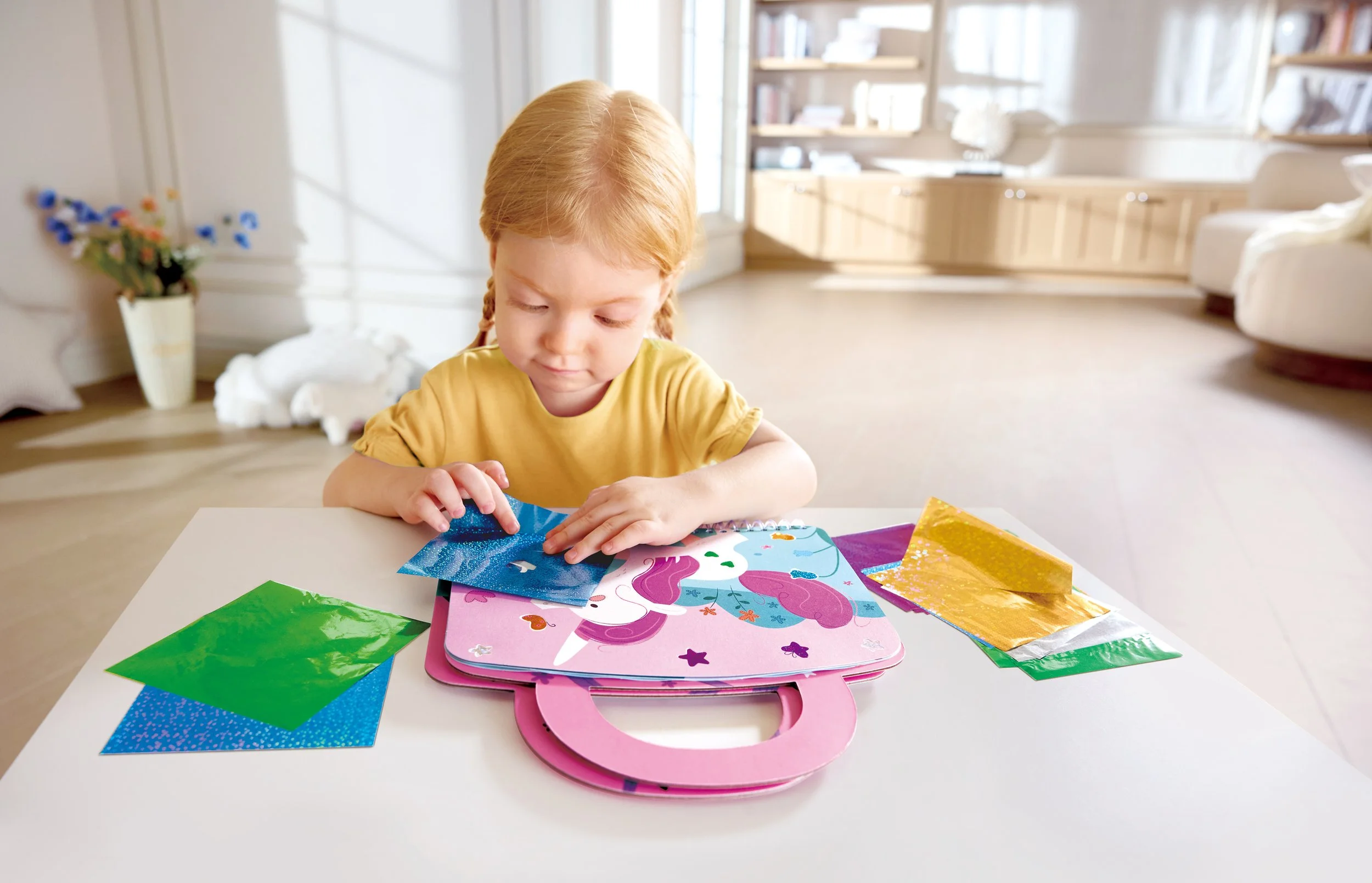 A young girl with red hair in braids, wearing a yellow shirt, plays with colored glossy paper and a unicorn-themed briefcase on a white table in a bright room.