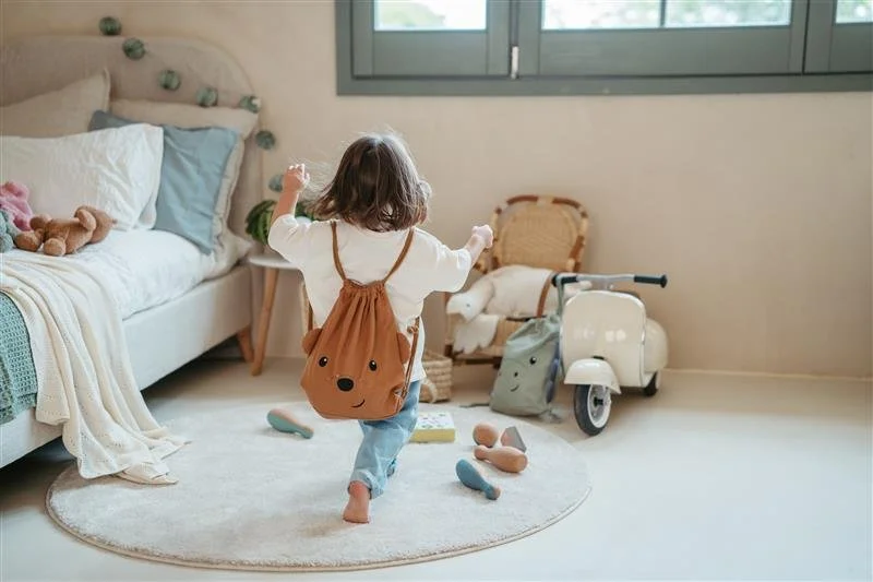 Child running in a cozy bedroom with a stuffed animal on the bed, a tricycle, and toys on the floor near a window.