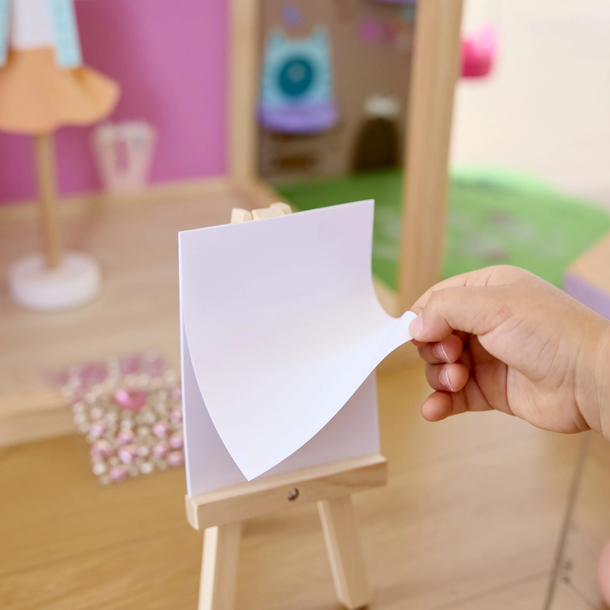 Person flipping a blank sheet of white paper on small wooden easel near pink, purple, and green background display.