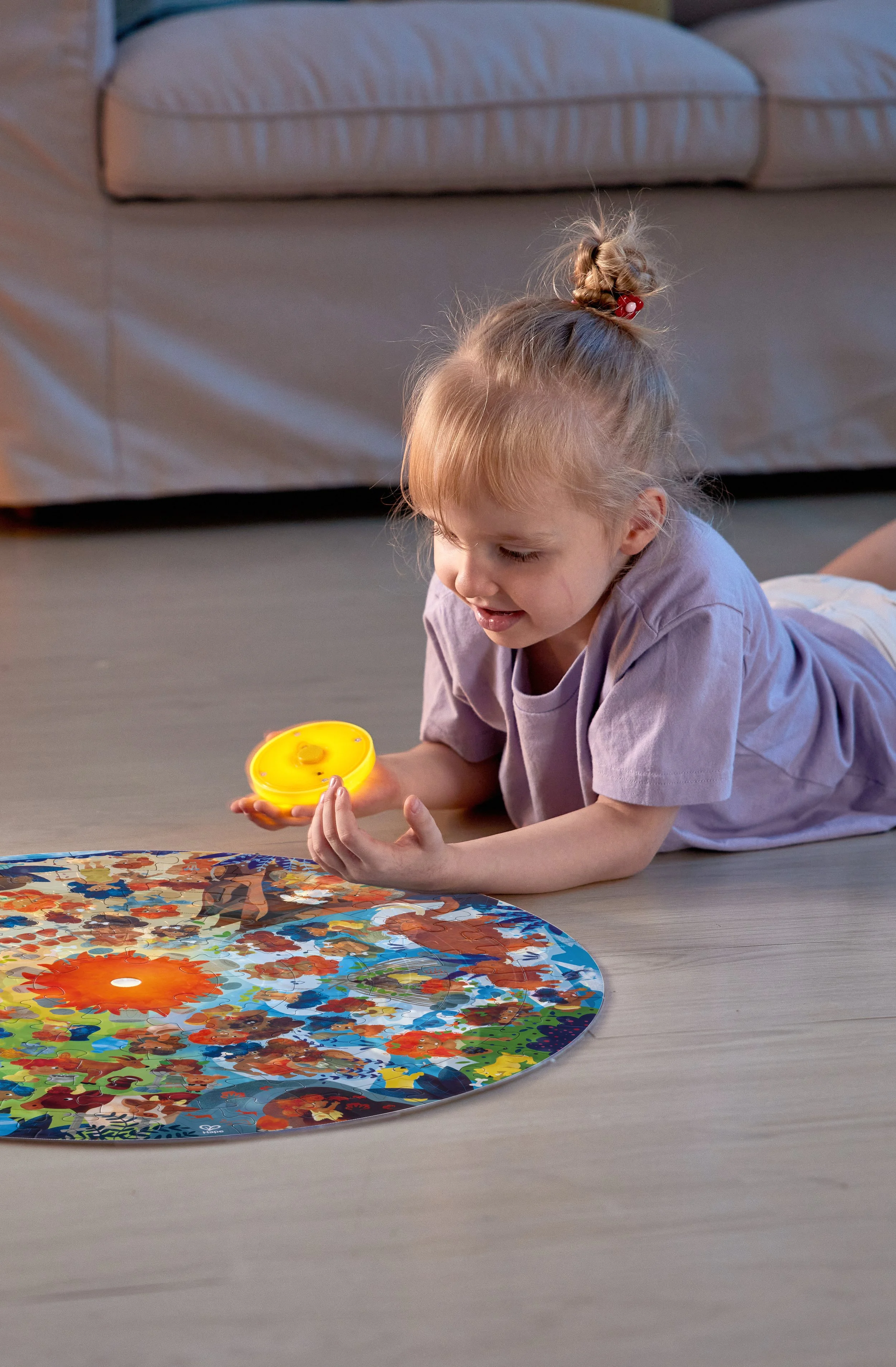 A young girl with blonde hair in a bun playing a colorful Hape puzzle on the floor, holding a yellow game piece.