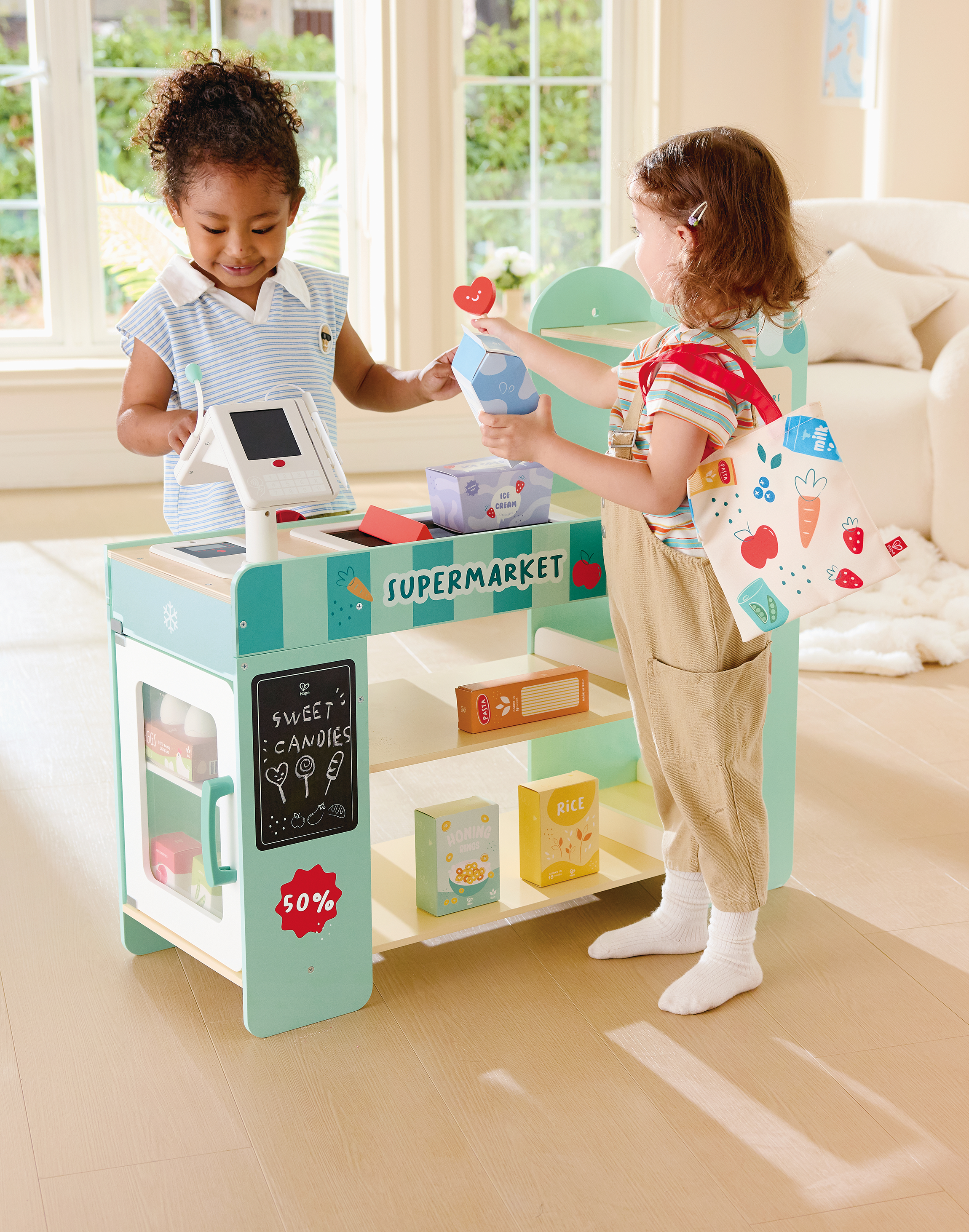 Two young girls playing pretend grocery checkout at a toy supermarket, with one girl checking out and the other handing her money, inside a bright, naturally lit room.