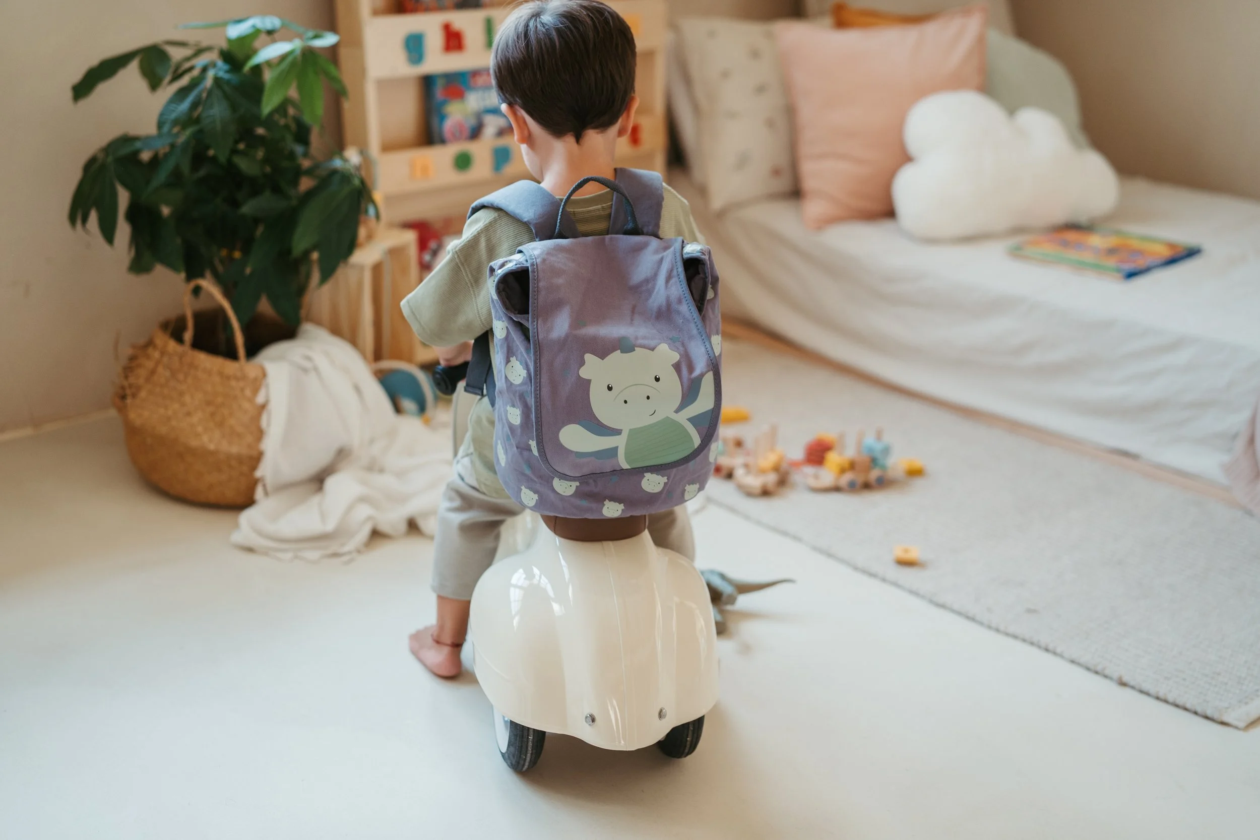 A young boy riding a white ride-on toy in a child's bedroom. He has a purple backpack with a cartoon animal on it. The room has a bed with pillows and a blanket, toys on the floor, a large plant in a wicker basket, and a bookshelf with children's boo