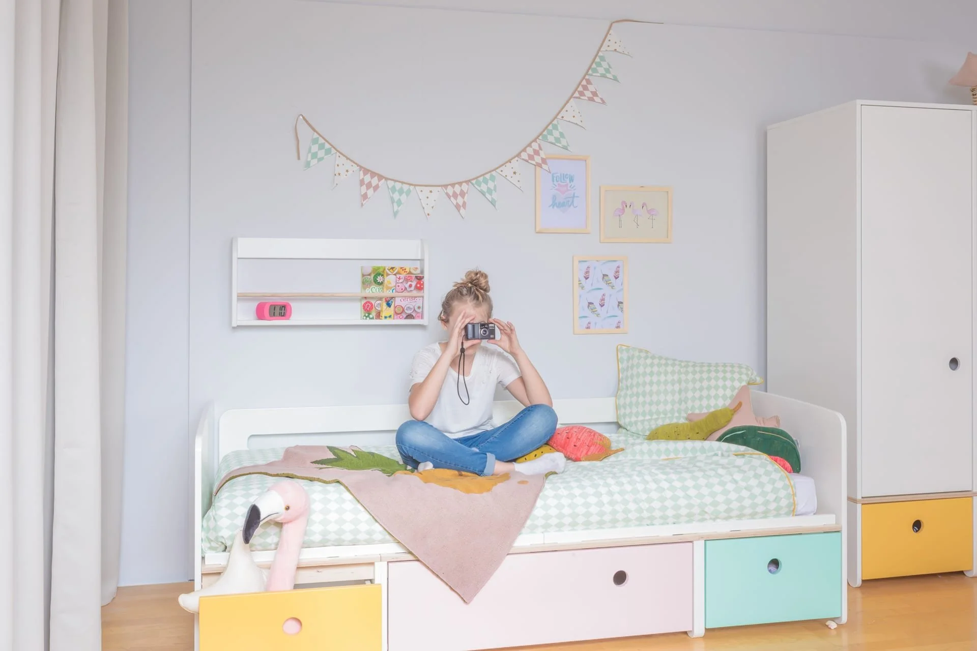 Young girl sitting cross-legged on a Colorflex bed with colorful cushions, holding a camera to her face, in a decorated child's bedroom with wall art, bunting, and stuffed animal