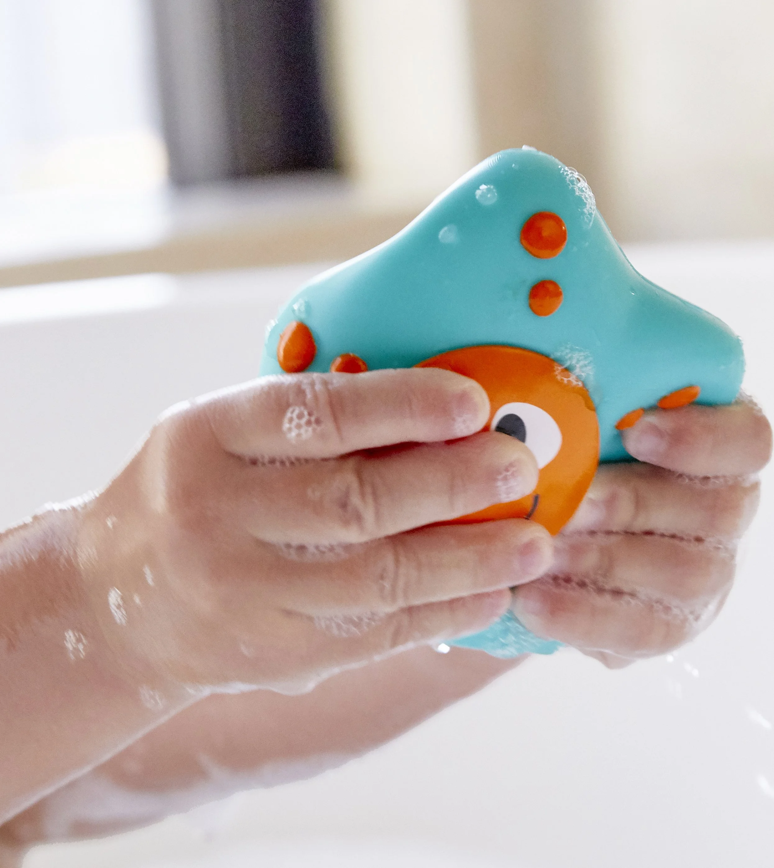 Child's hand holding and playing an Hape bath toy from the Ocean floor squirter" set, covered in soapy bubbles during a bath.