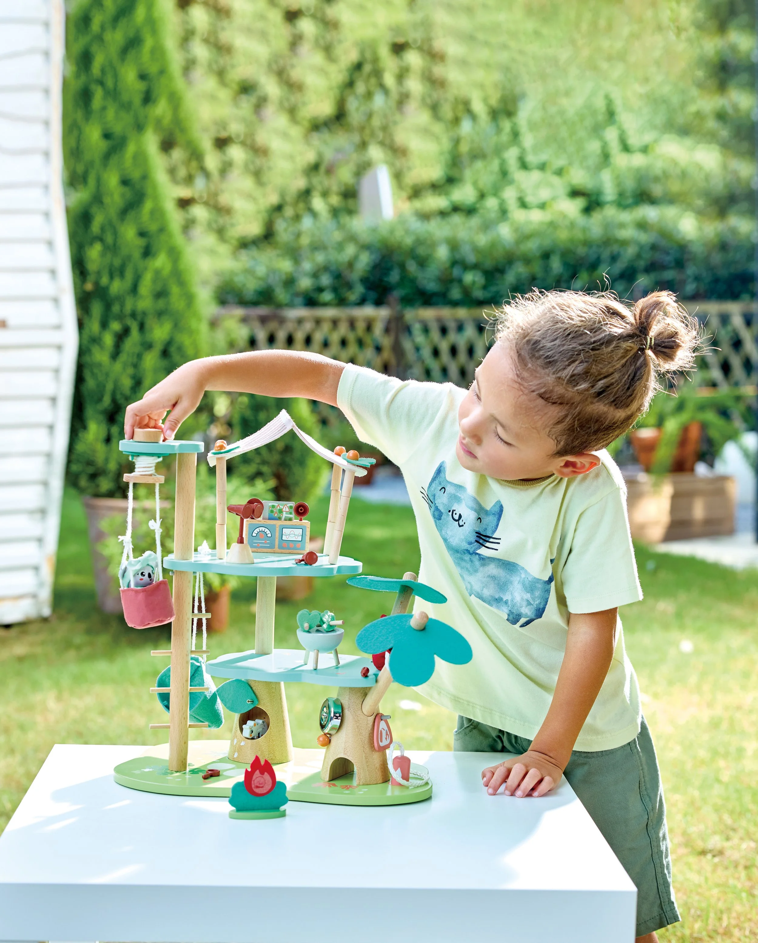 Young boy playing with the Green Planet Explorers Forest Keepers Headquarter by Hape, a wooden toy fire station, with greenery and a wooden fence in the background.