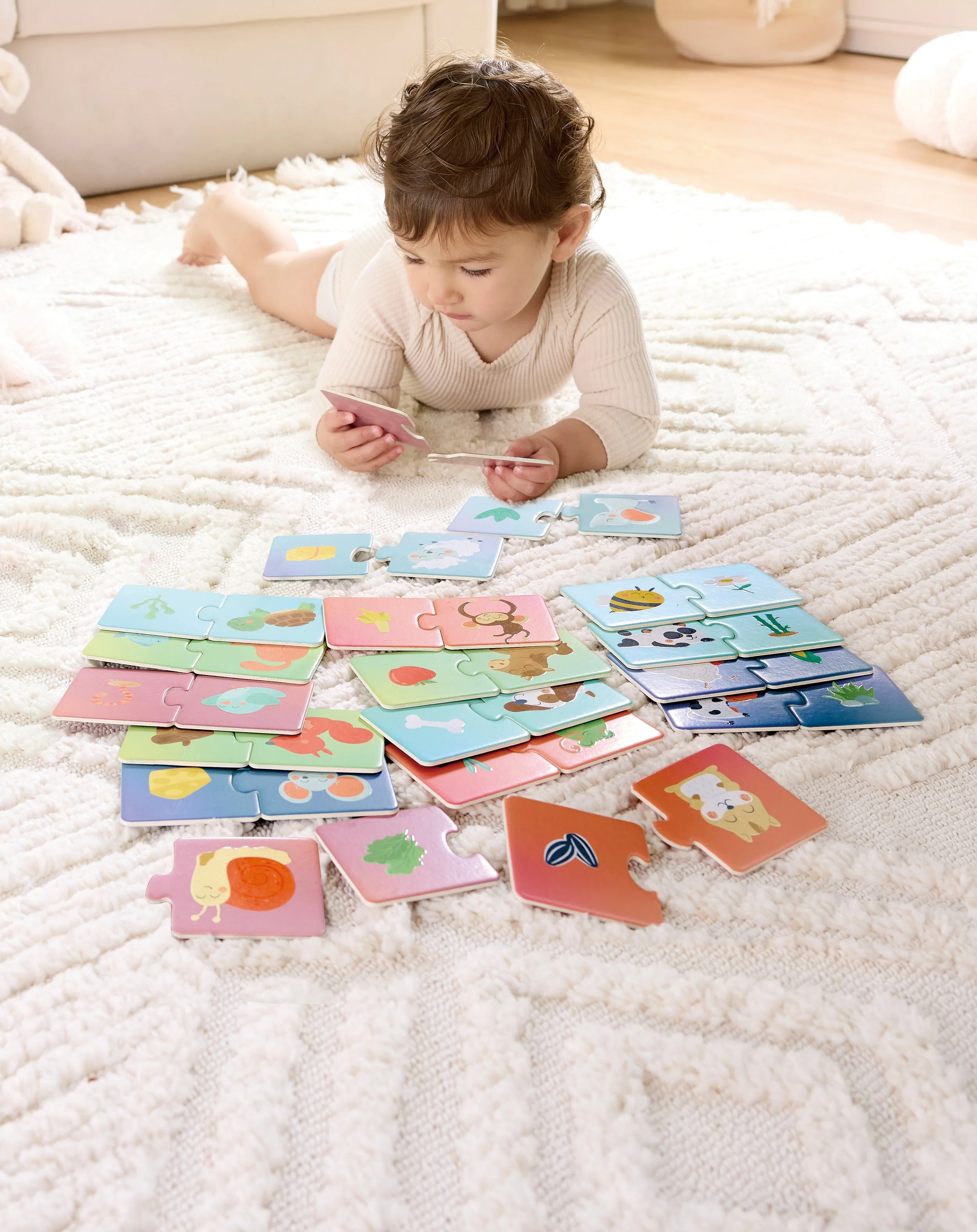 A young child lying on a plush white rug playing with colorful animal and nature-themed Hape jigsaw puzzle pieces.