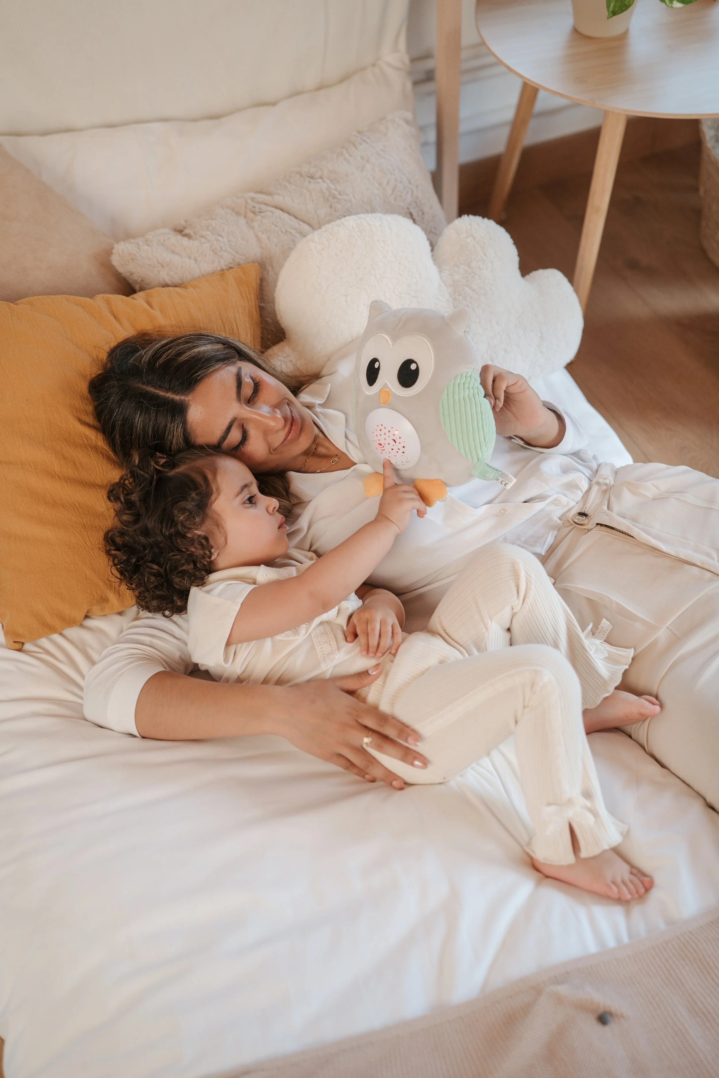 A woman and a young girl are lying on a bed, with the girl reaching out to touch a Archi plush projector from Eurekakids Cucu held by the mum.