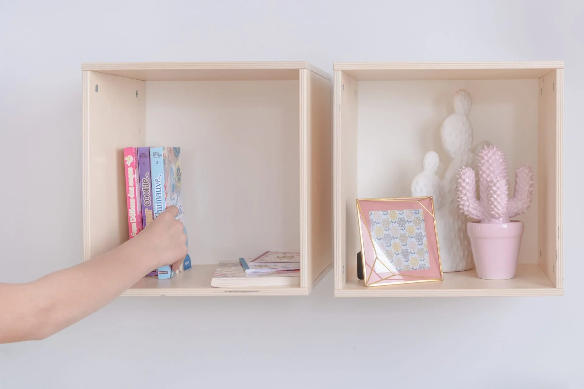 A child reaching to select a book from a light wooden wall shelf with two sections by Colorflex, containing books and decorative items.