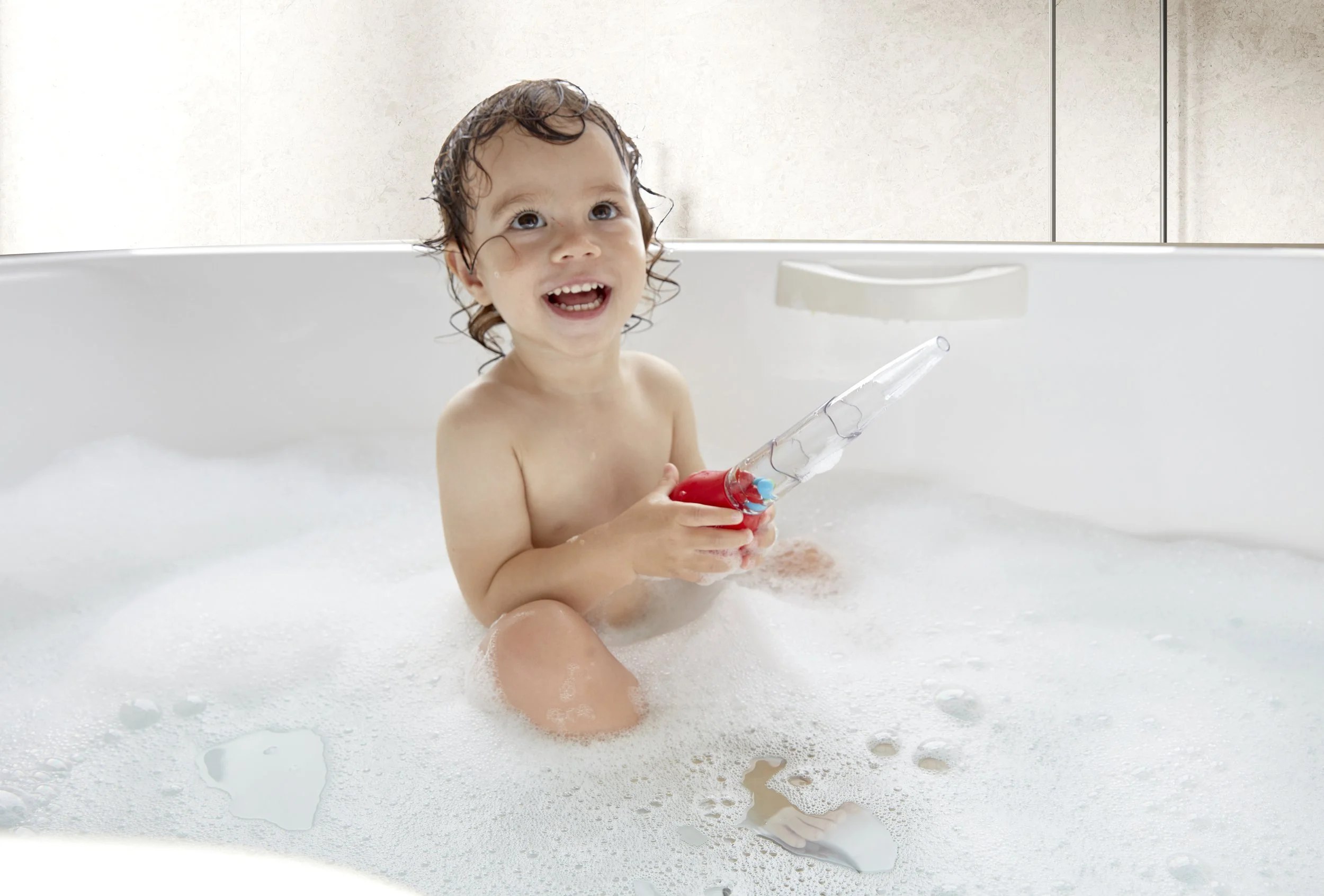 A young child with wet hair smiling in a bubble-filled bathtub holding "Squeeze and squirt pippette" by Hape.