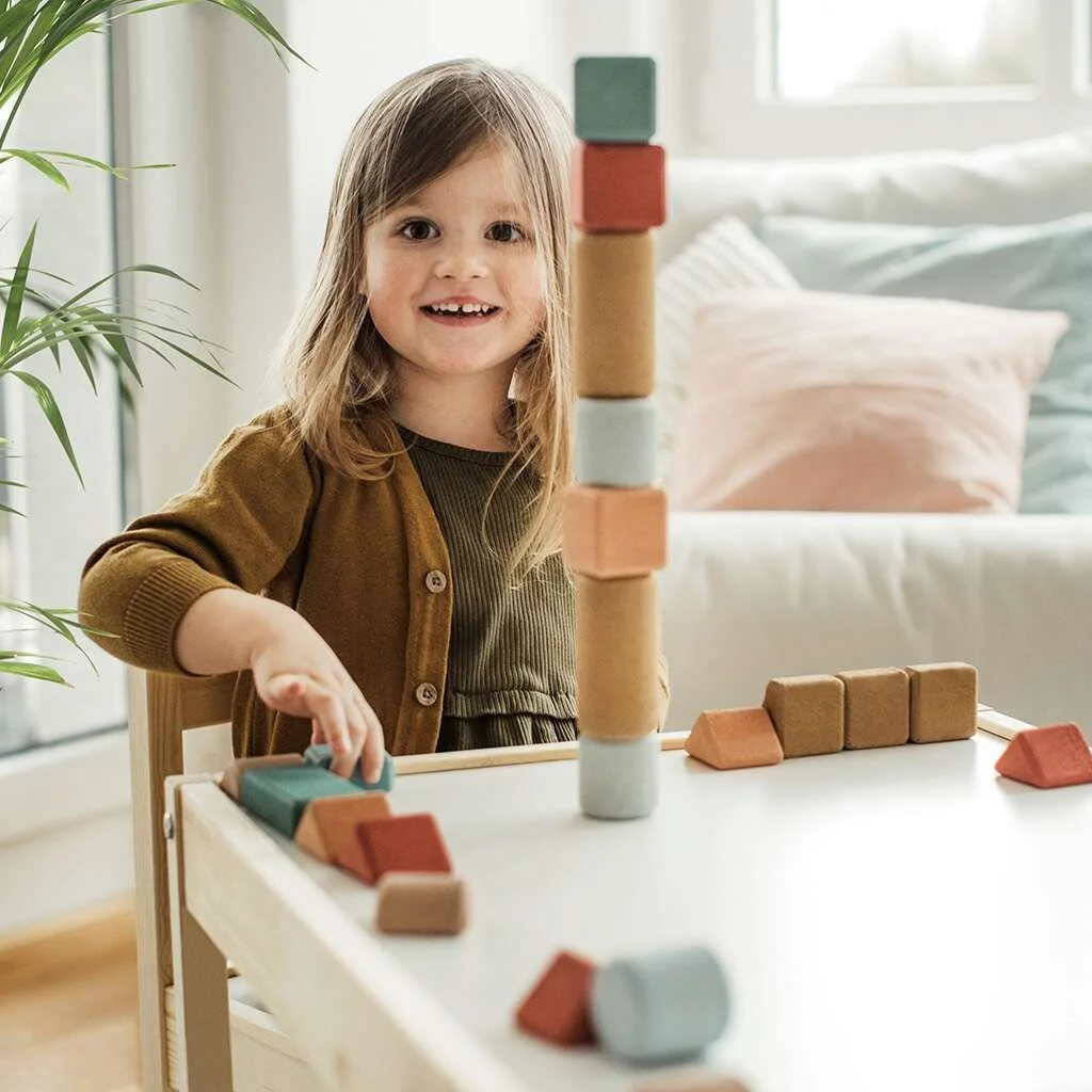 A young girl with long hair and a brown cardigan smiling while playing with colorful cork blocks on a table in a bright room. By Korko.