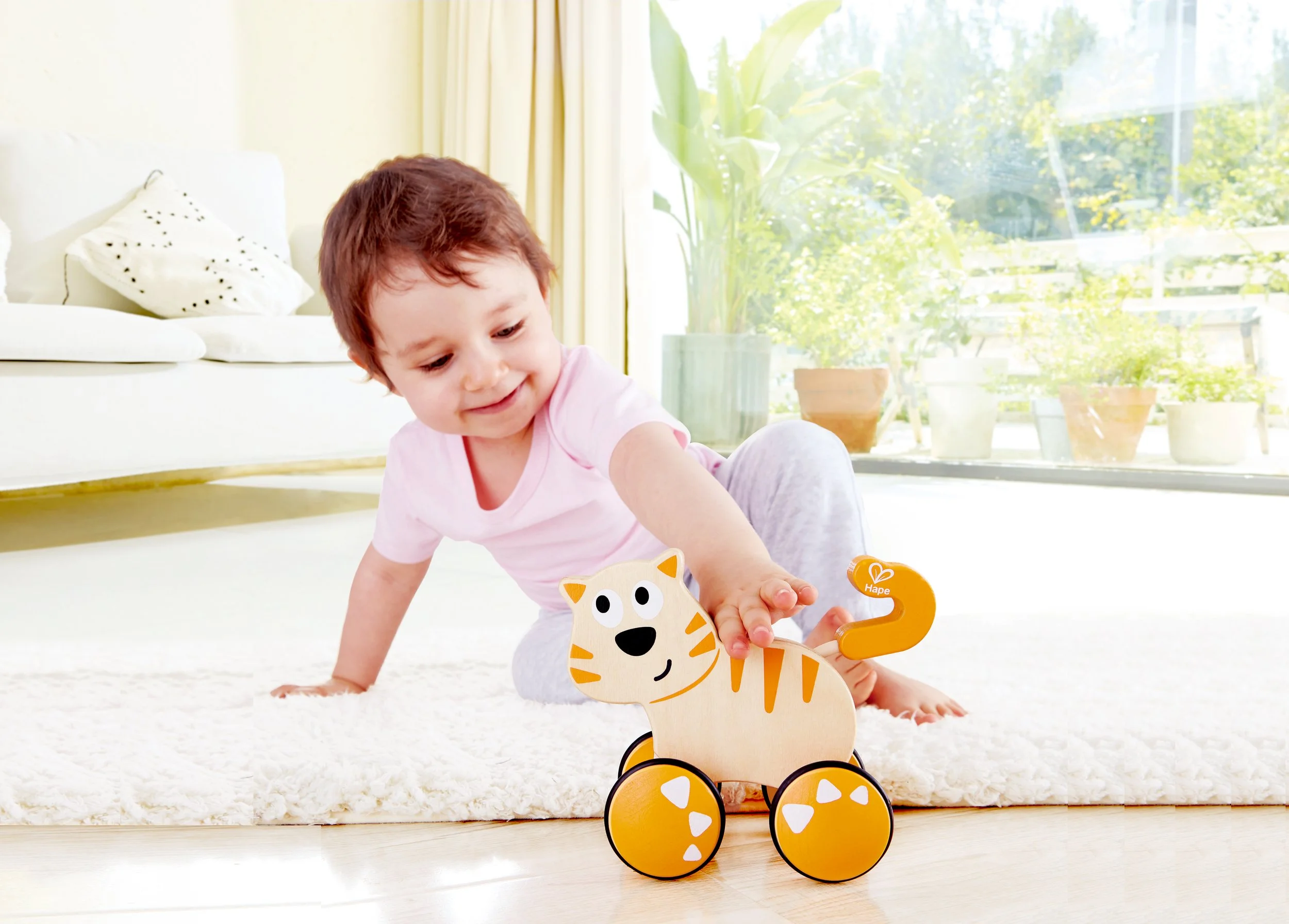 A young child playing with a wooden toy cat on a white carpet in a bright living room with large windows, plants, and a cream-colored sofa. Developed by Hape.