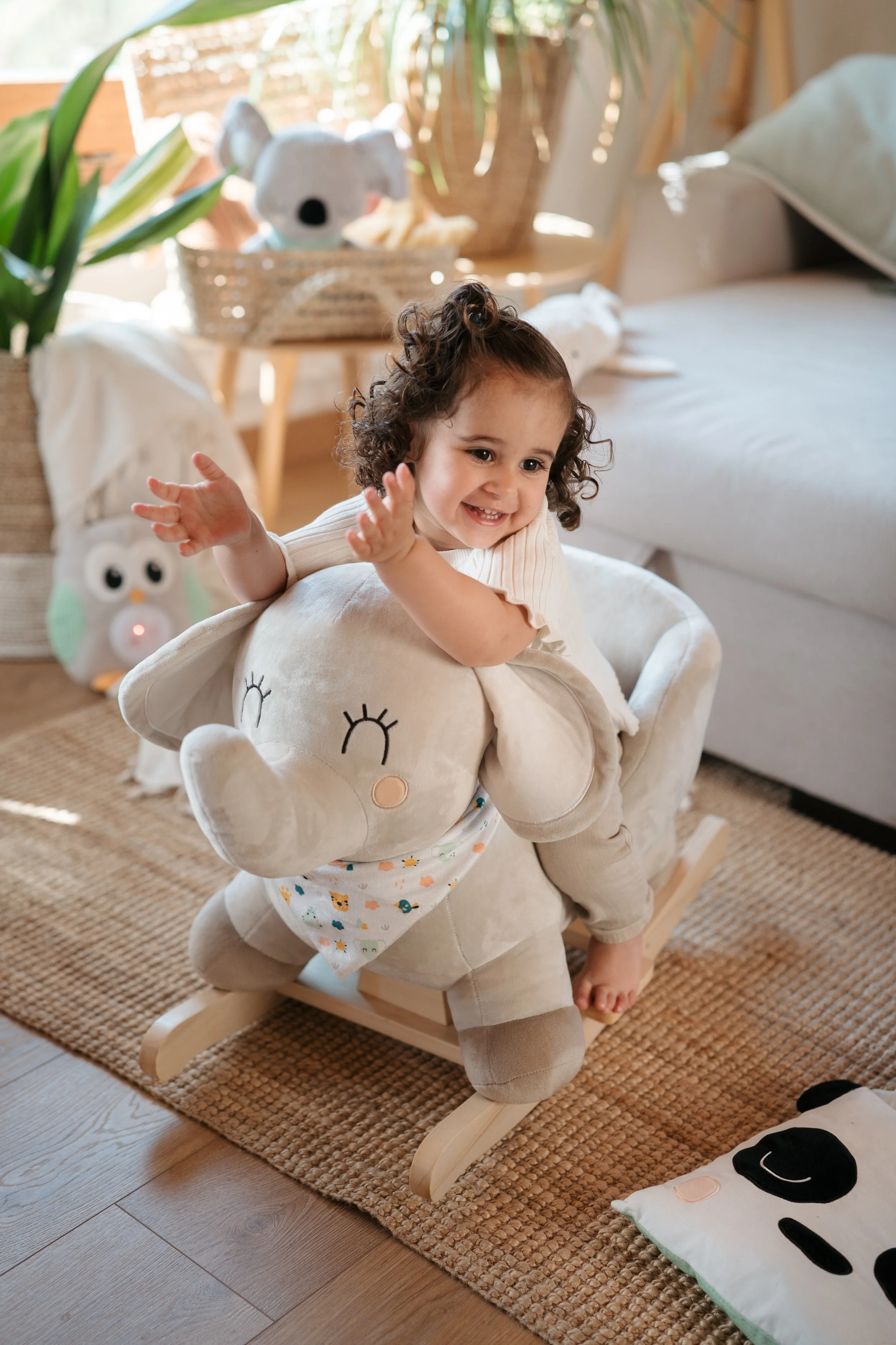 A young girl with curly hair sitting on a plush rocking elephant by Eurekakids Cucu, in a cozy living room. The room features plants, stuffed animals, and natural light, creating a warm and playful atmosphere.