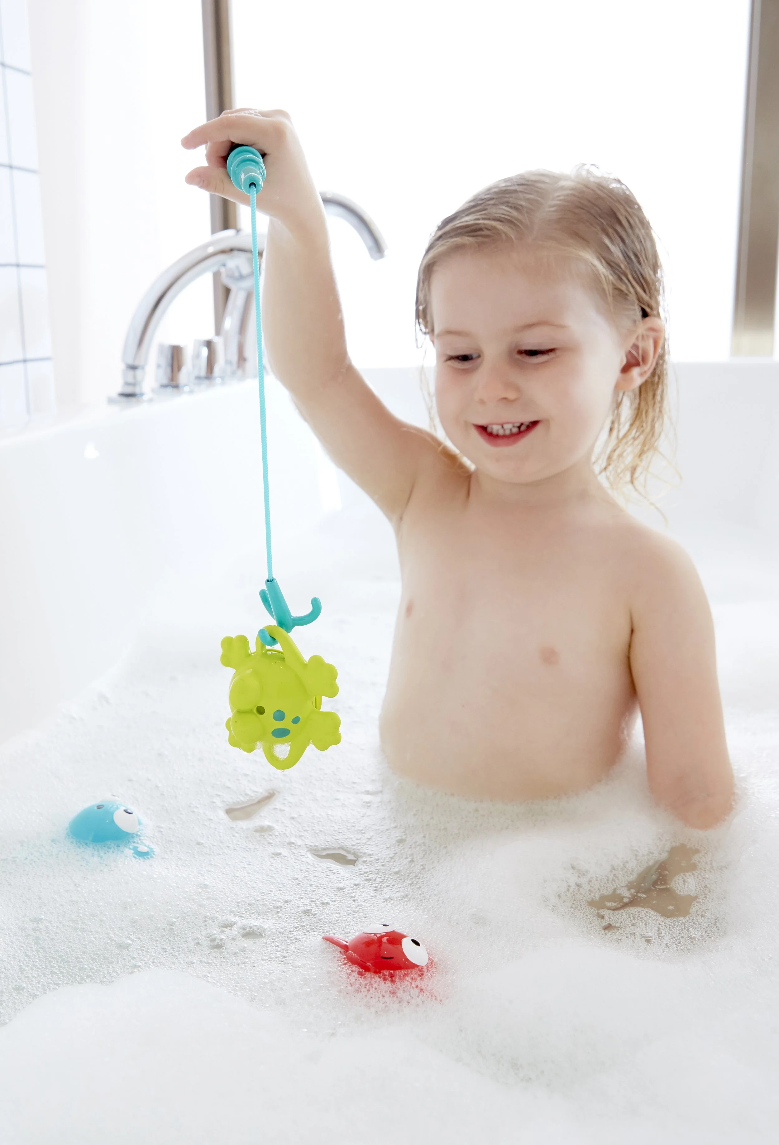 A young child with wet hair playing in a bubble-filled bathtub playing with "Double fun fishing set" by Hape.